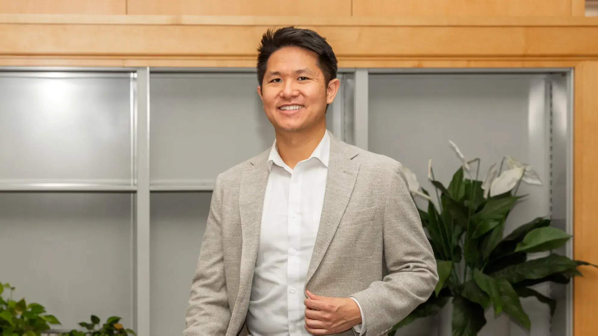 A person smiling while standing indoors, wearing a light grey suit jacket and white shirt.