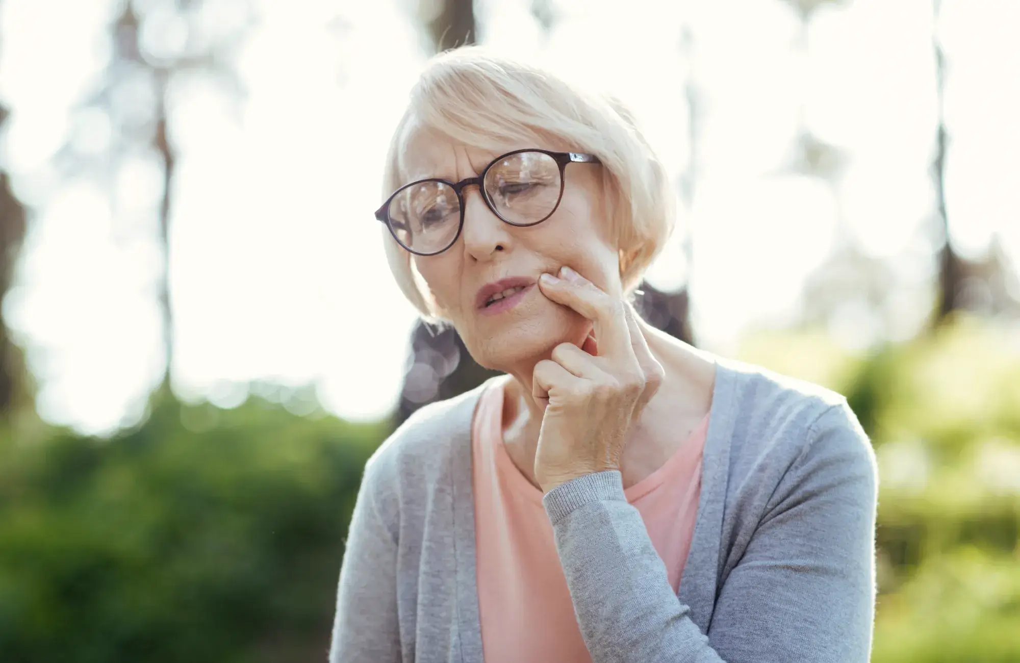 An elderly woman touches her cheek, appearing to be in pain or discomfort.