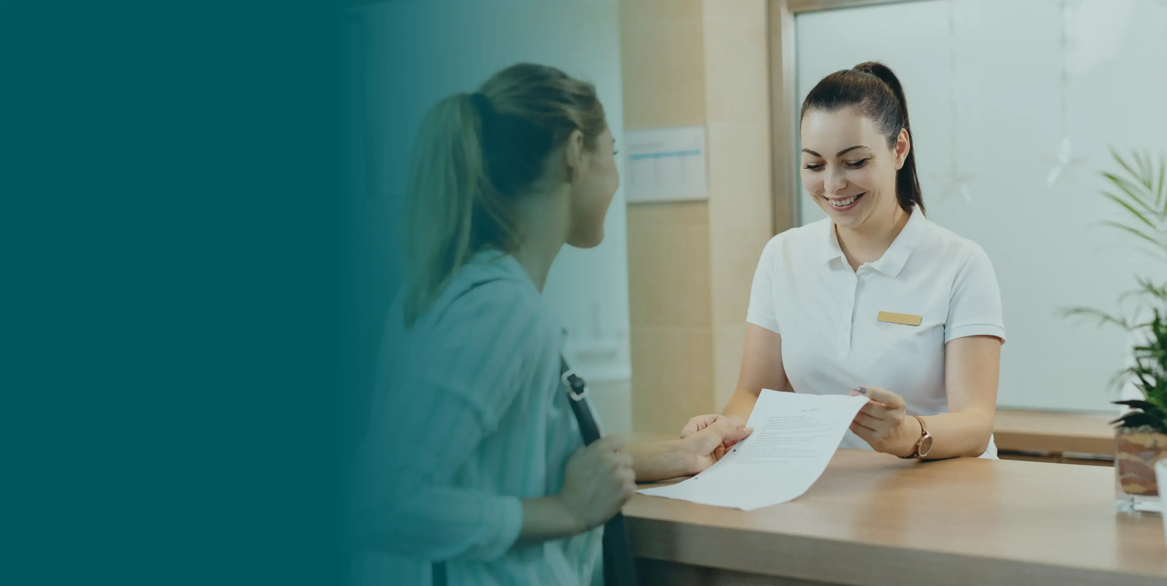 Two women talk at a reception desk; one holds a document, the other listens attentively.