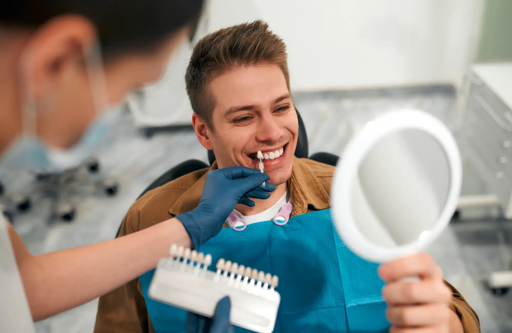 A dentist uses a shade guide on a smiling man's teeth as he checks with a hand mirror.