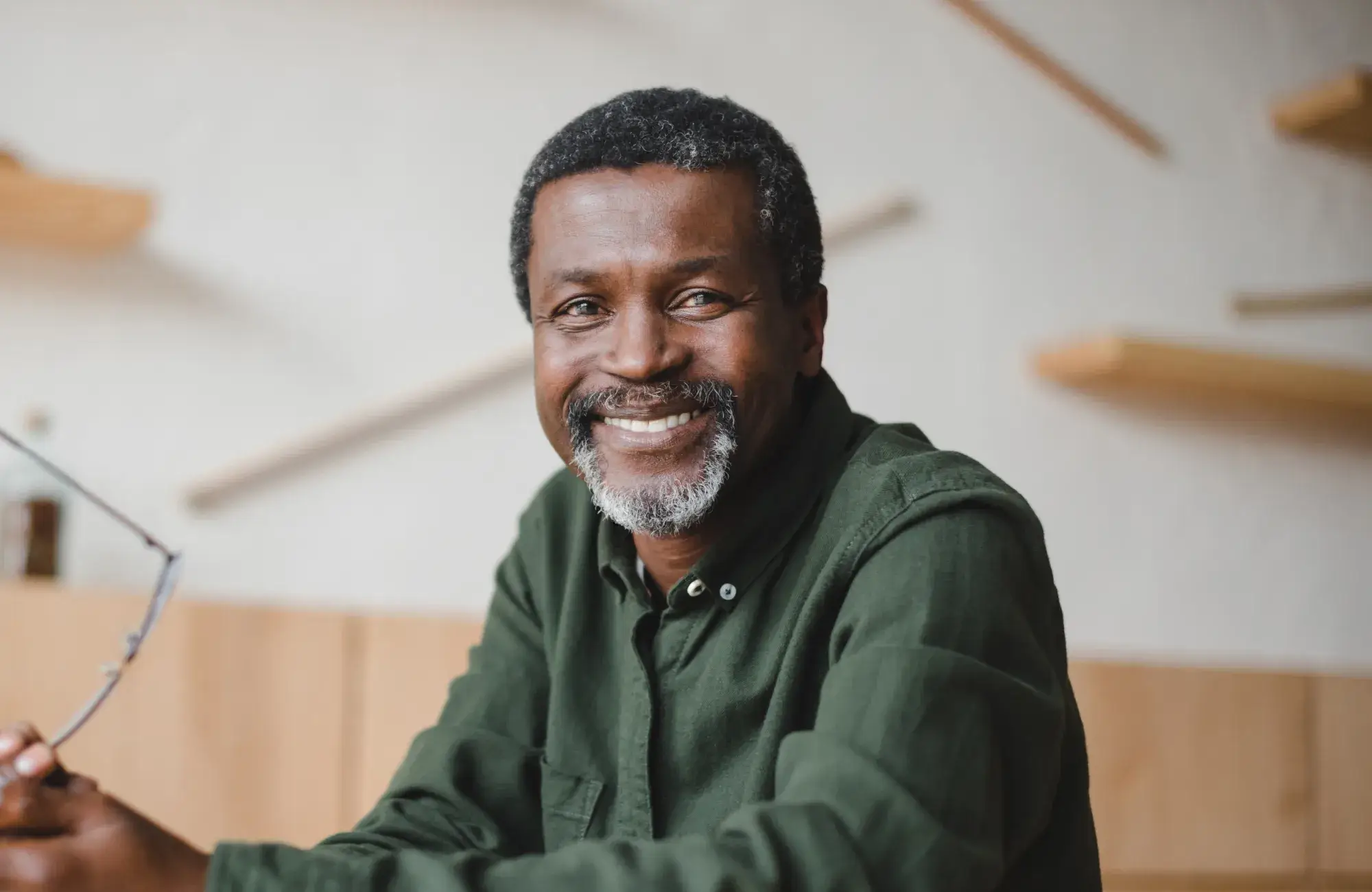 A smiling man in a green shirt sits holding glasses, looking at the camera.