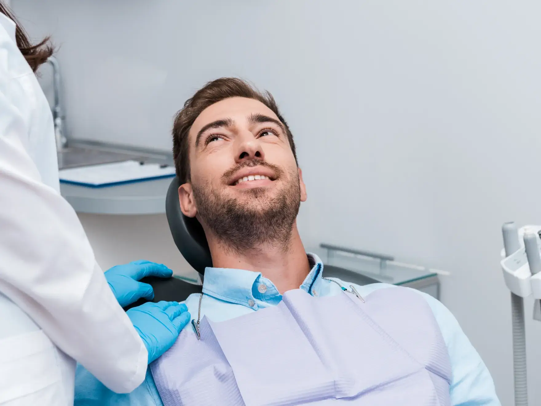 Man sitting in a dental chair, smiling up at a dentist with gloved hands on his shoulder.
