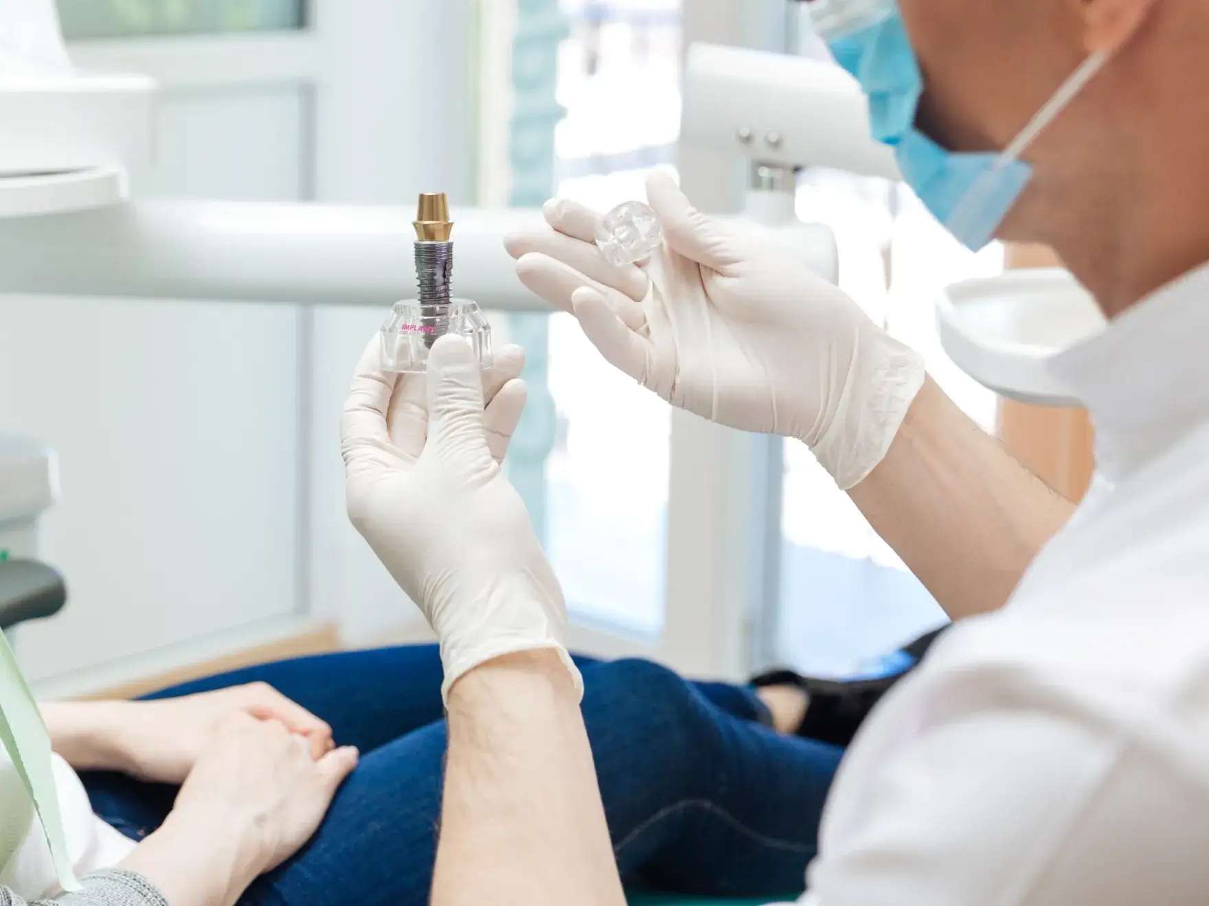 A dentist in gloves and mask holds a dental implant model, explaining its use to a patient.