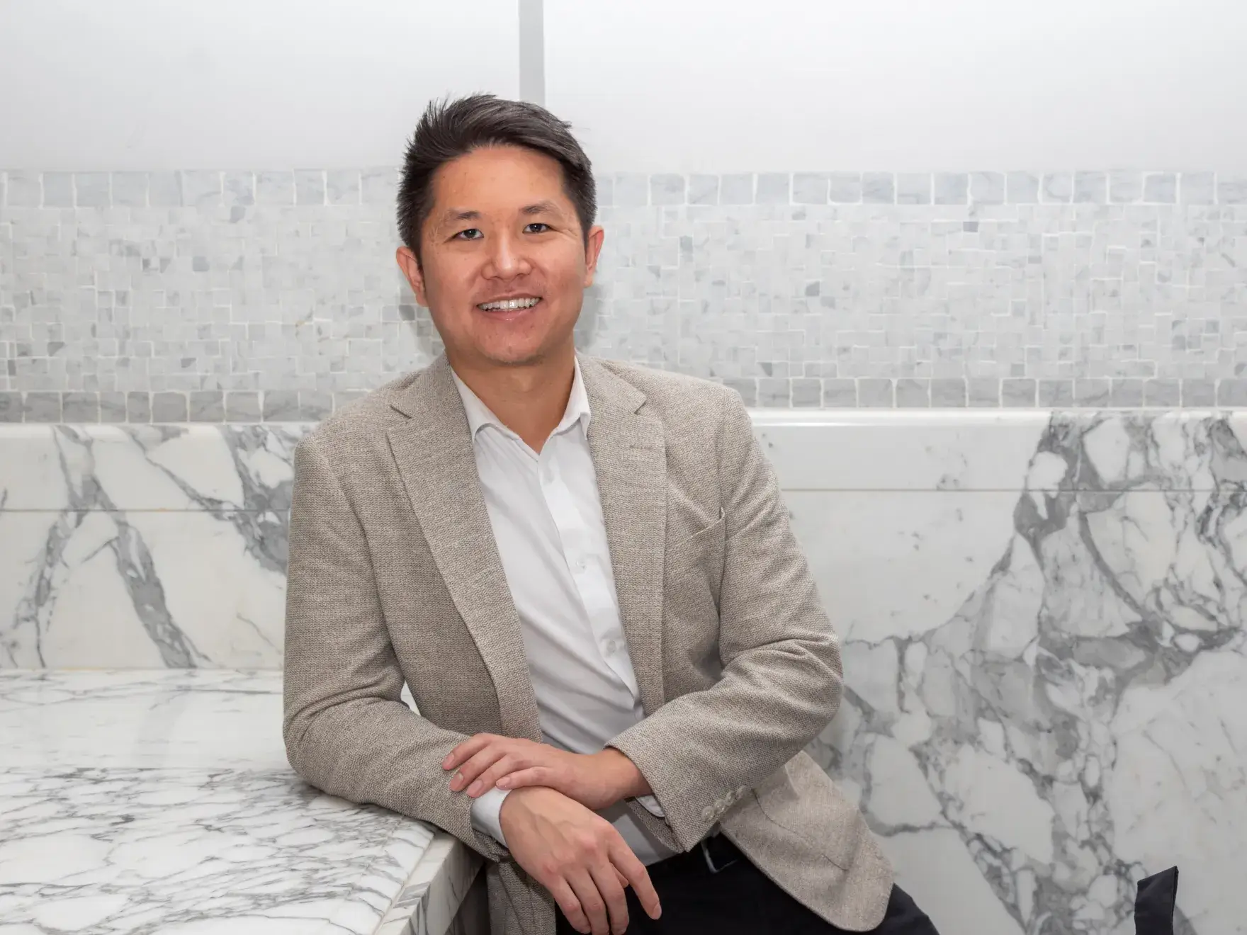 A person in a beige blazer is leaning against a marble counter, smiling at the camera.