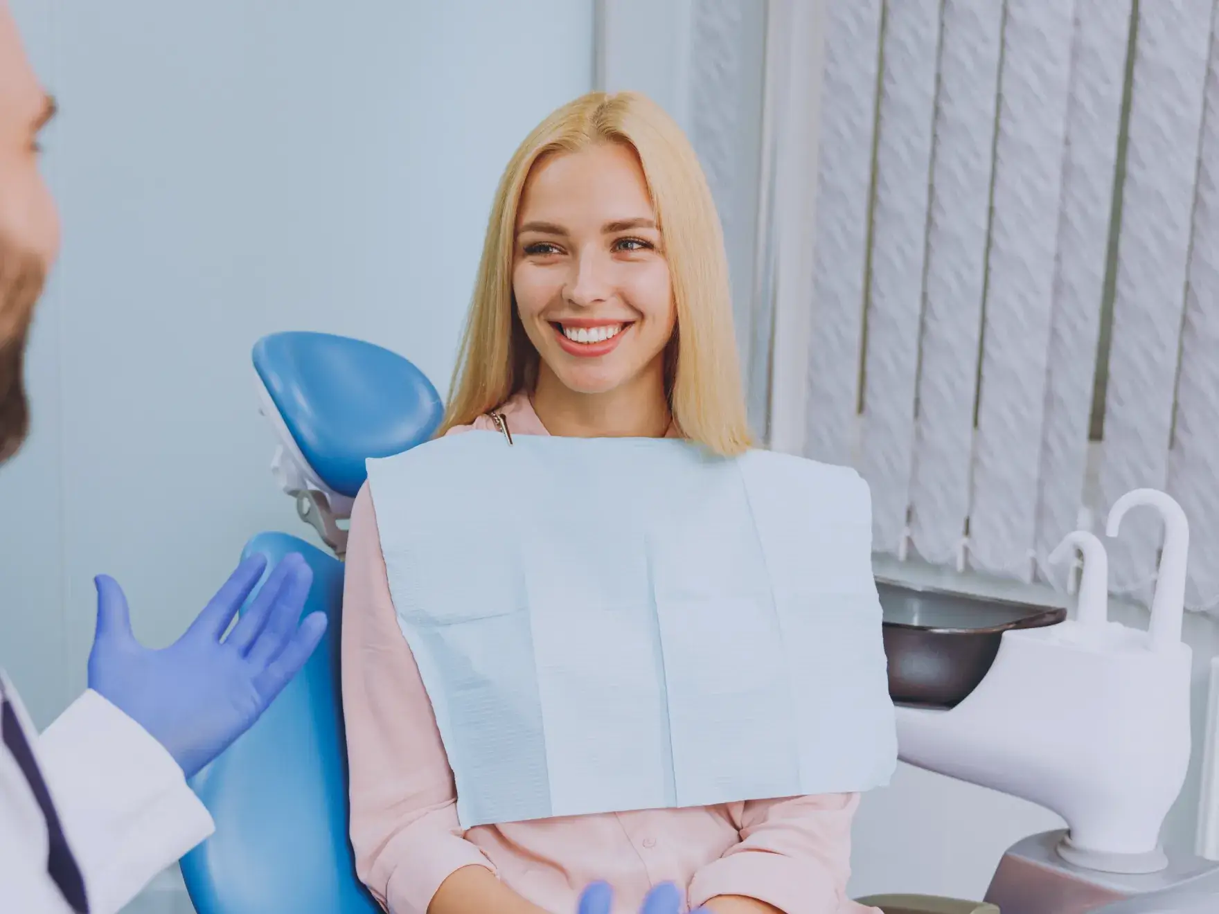 A woman sits in a dental chair, smiling at a dentist wearing gloves who is talking to her.