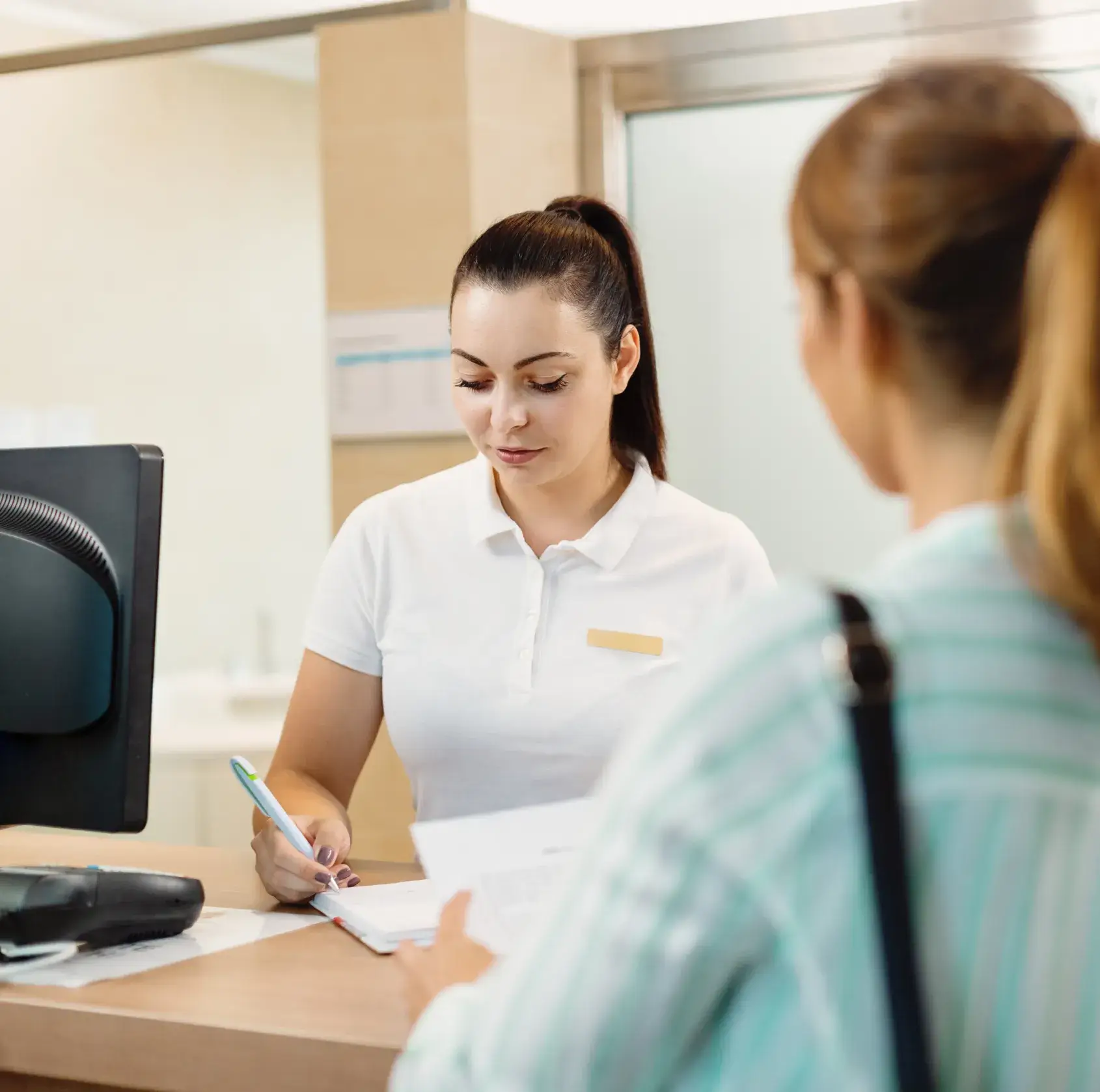 A receptionist in a white uniform helps a woman fill out a form at a desk.