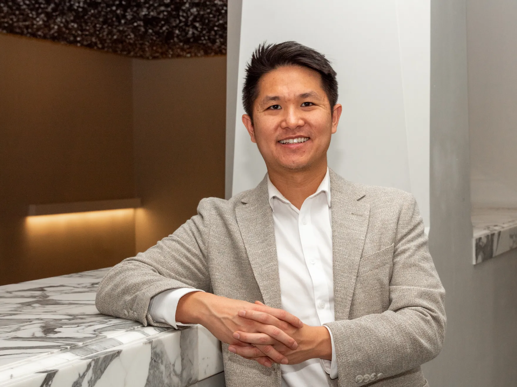 A man in a gray suit jacket stands with hands clasped, leaning on a marble surface.