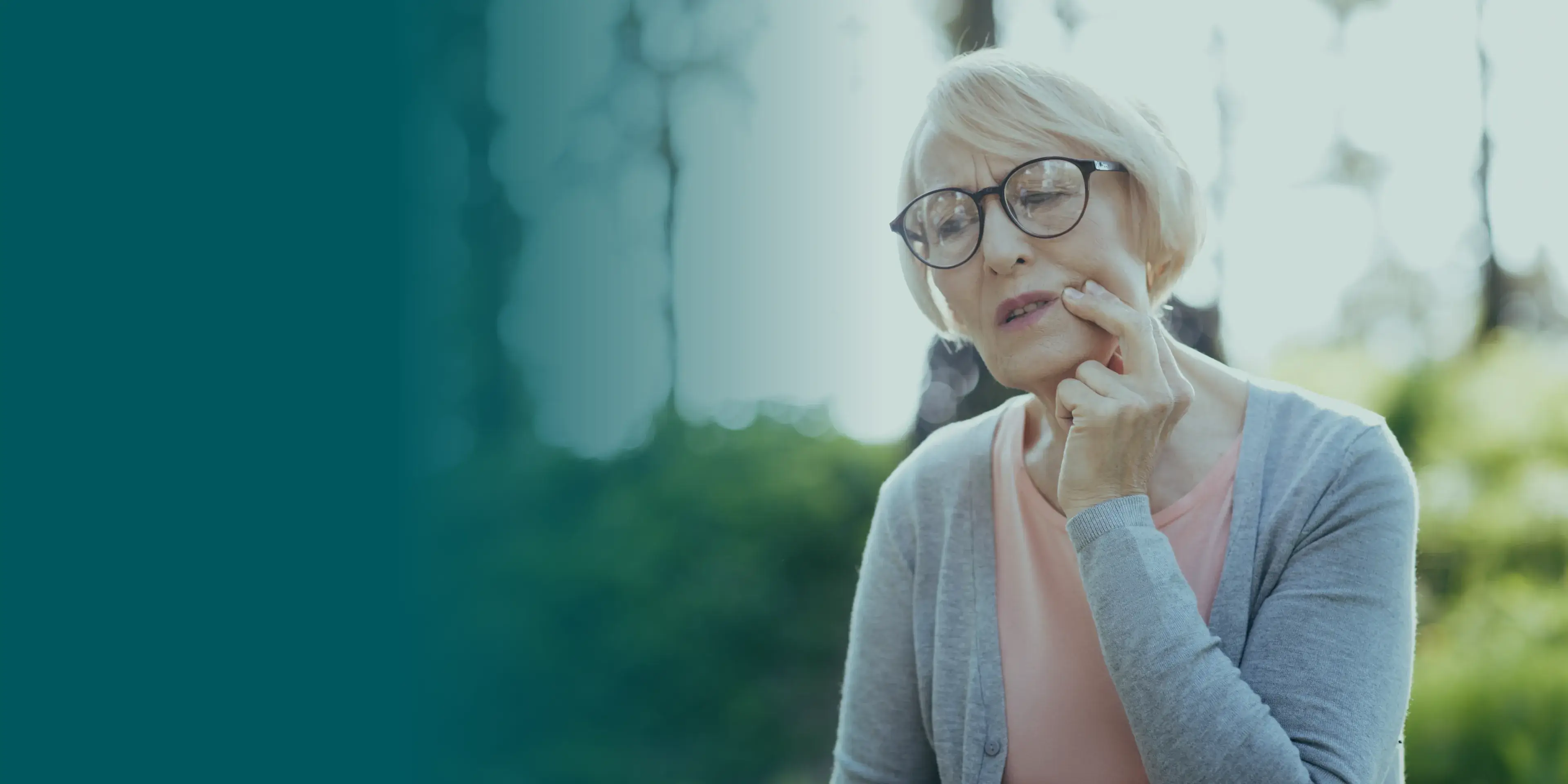 Elderly woman with glasses touching her cheek, sitting outdoors, appears thoughtful.