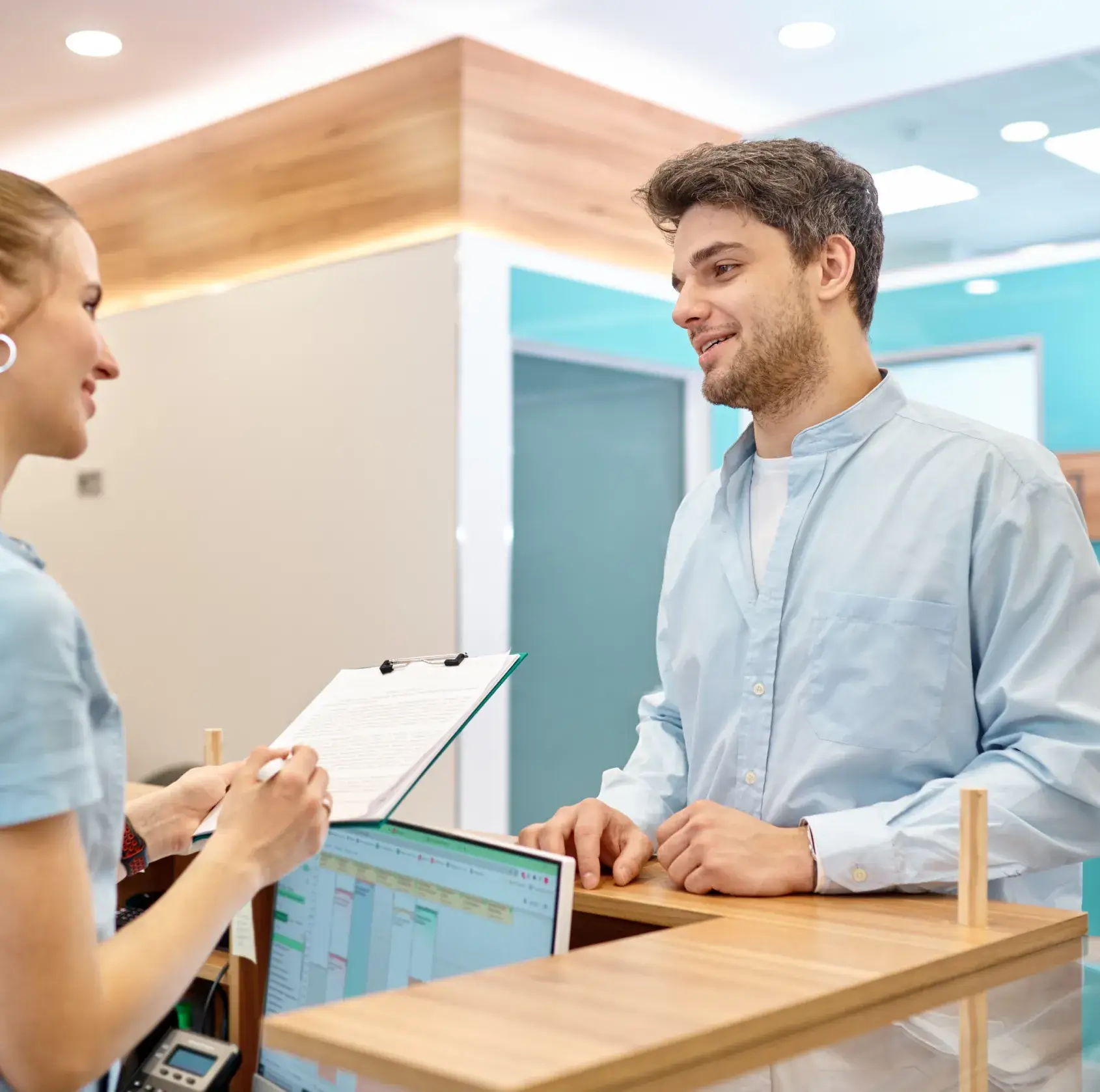 A man in a light blue shirt talks to a receptionist holding a clipboard at a desk.