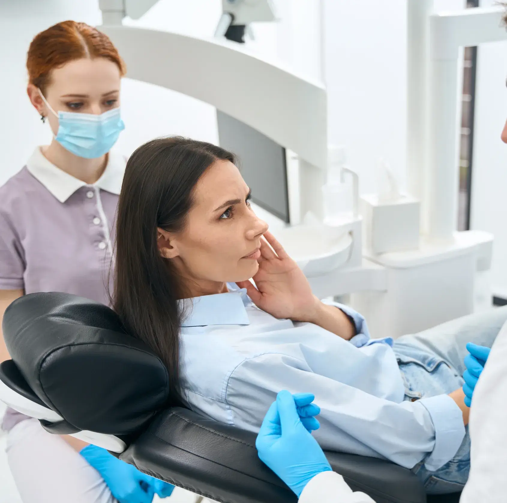 A woman sits in a dental chair, holding her jaw, while a dentist and assistant observe.