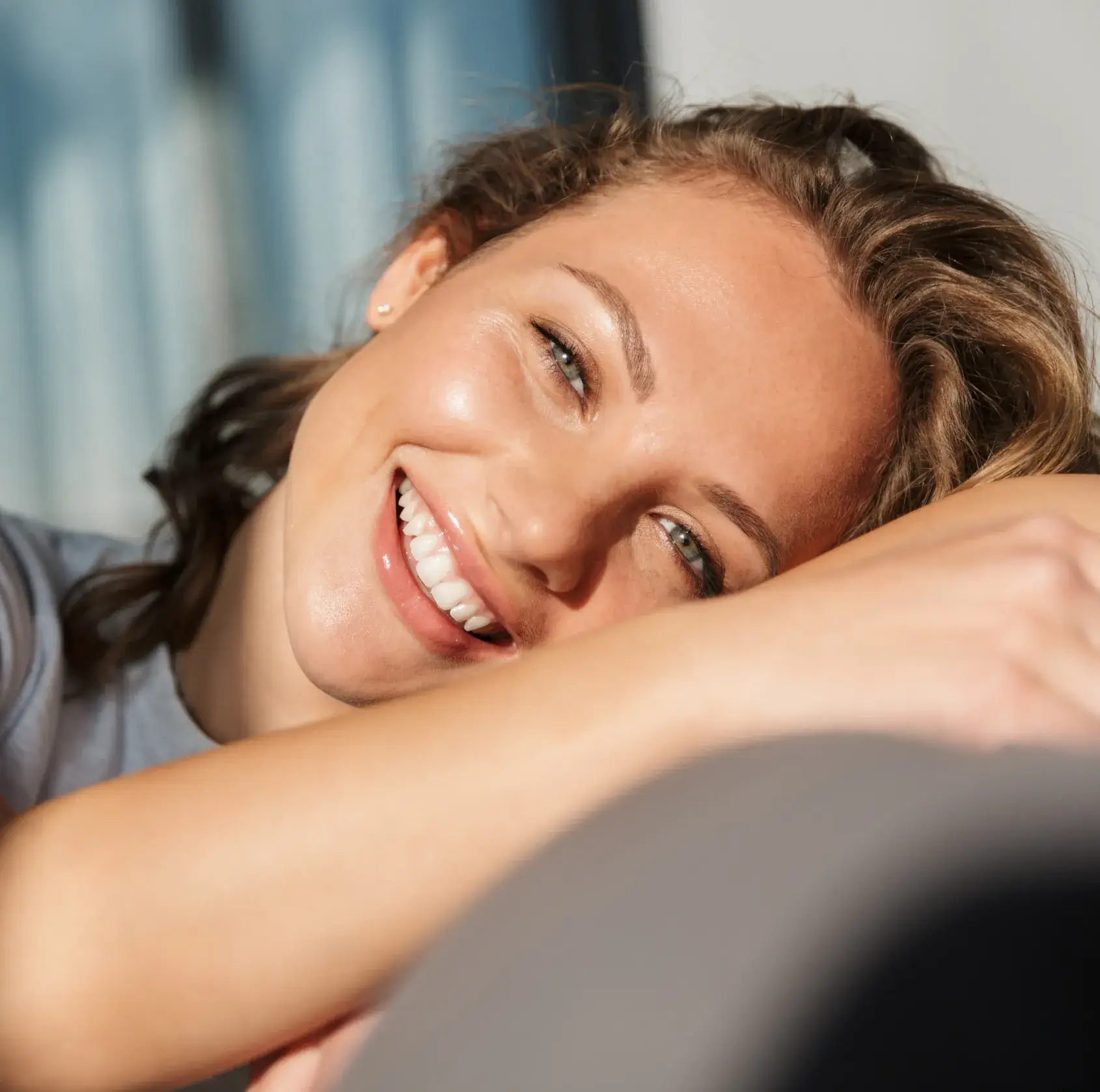 A person smiles brightly, resting their head on their arm while looking at the camera.