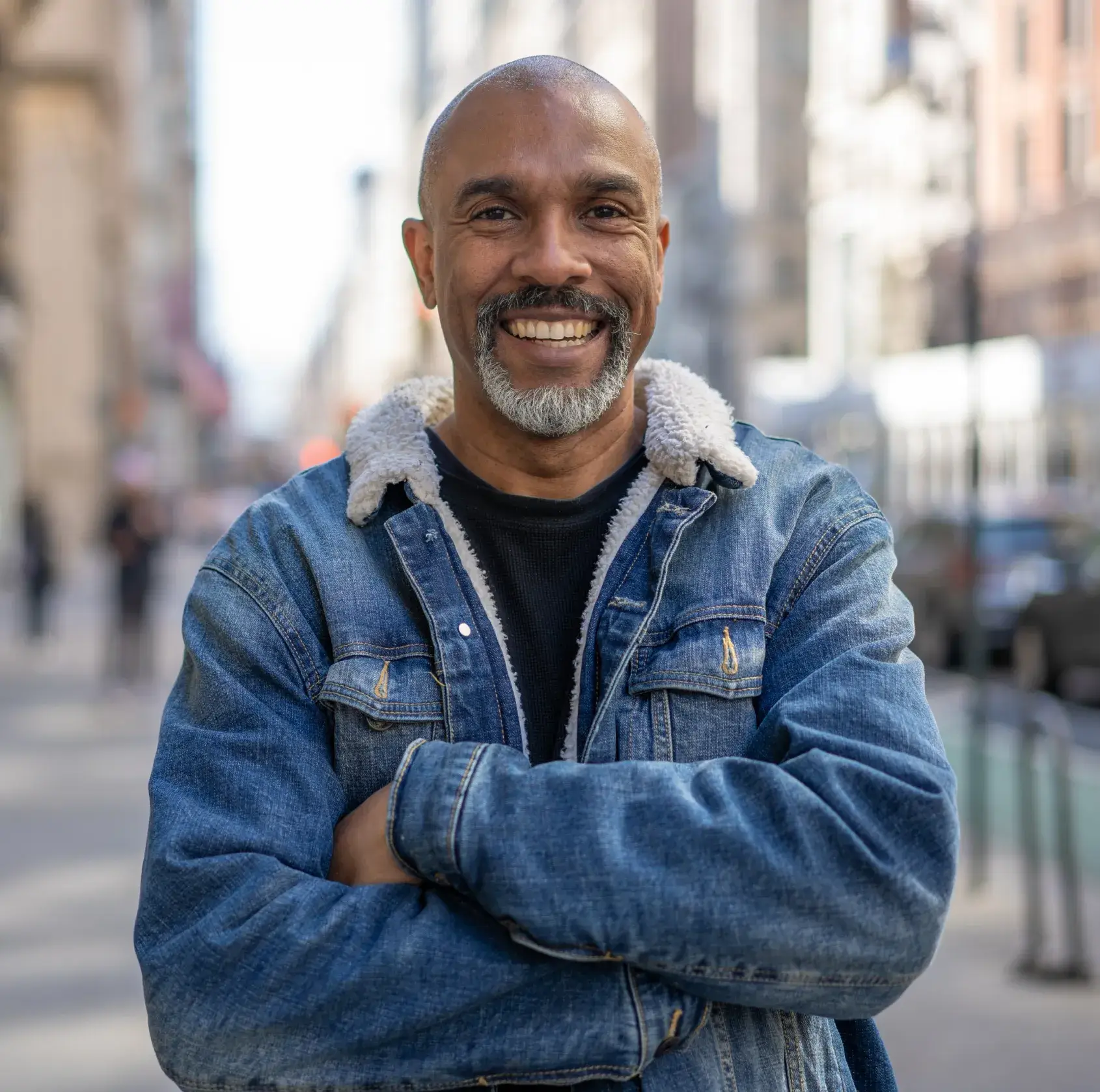 A man stands on a city street, smiling with his arms crossed, wearing a denim jacket.