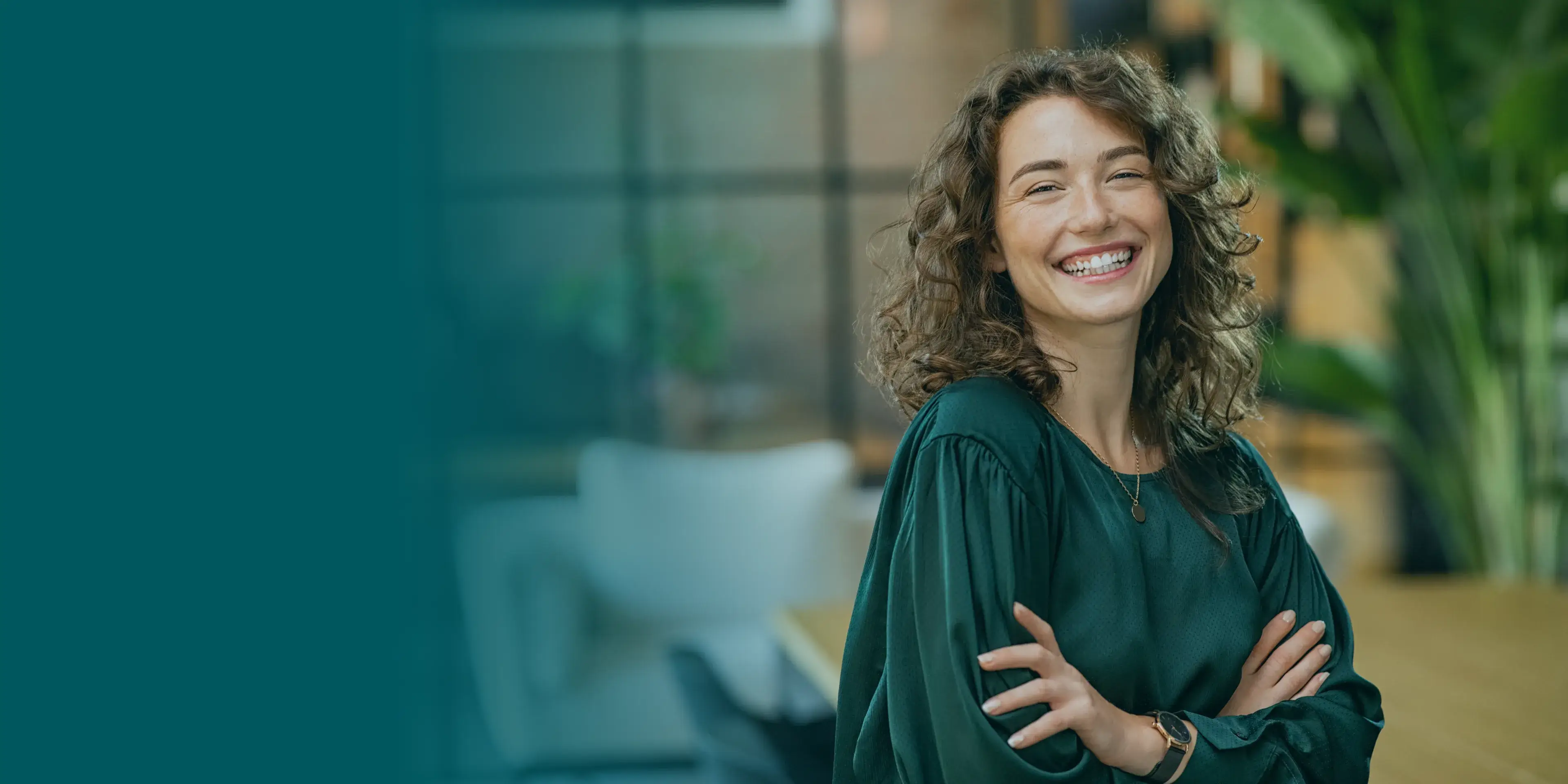 Smiling person with curly hair stands with arms crossed in an office setting.