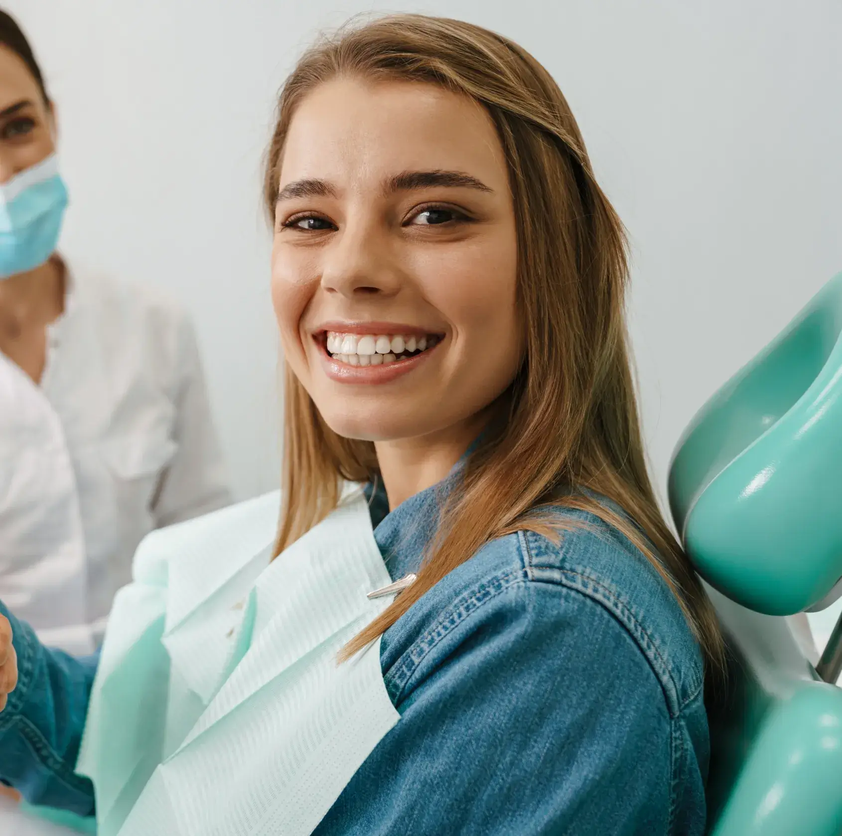 A woman sits in a dental chair, smiling, with a dentist nearby wearing a mask.