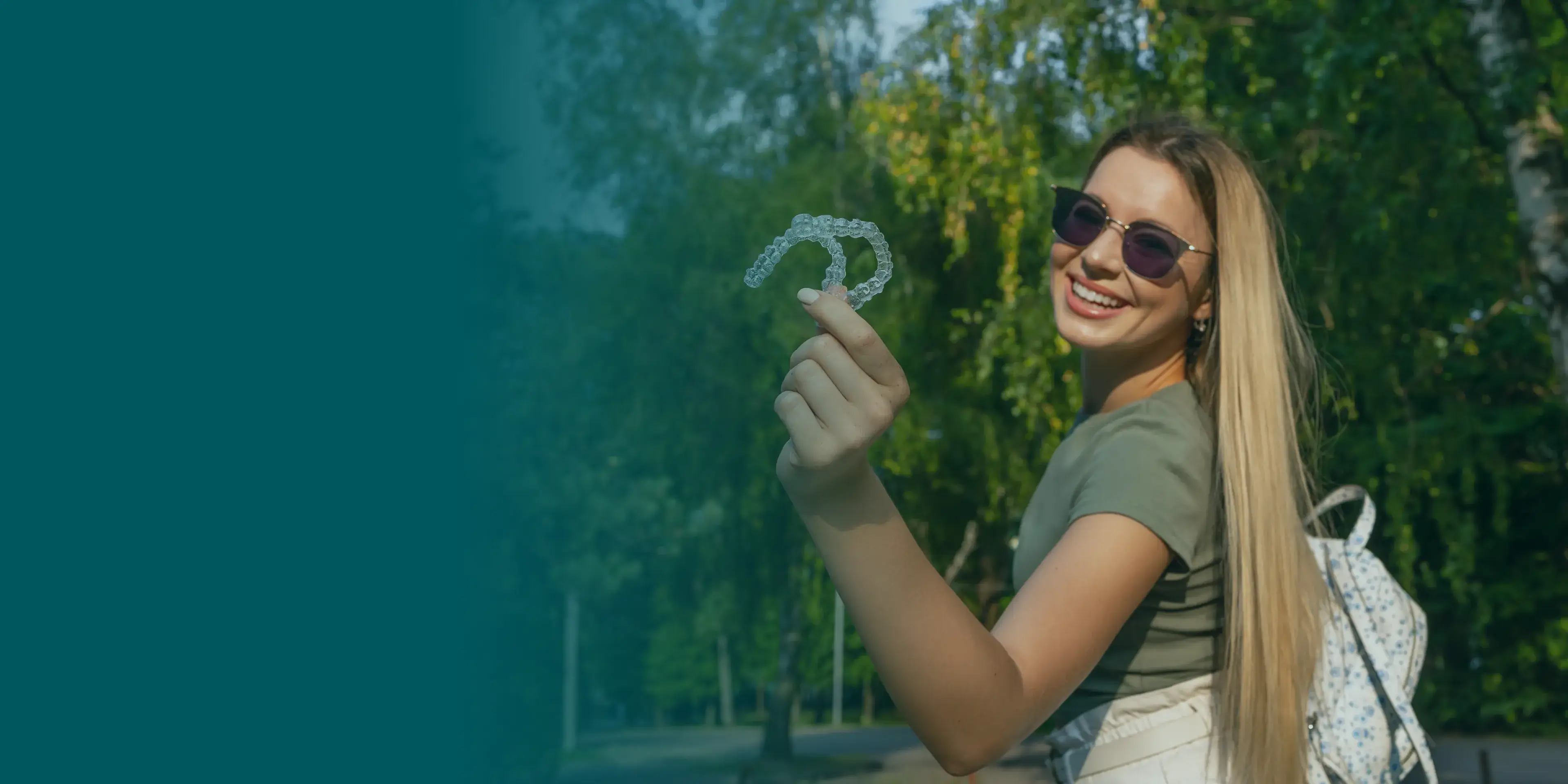 A woman wearing sunglasses smiles and holds a clear dental aligner outdoors.