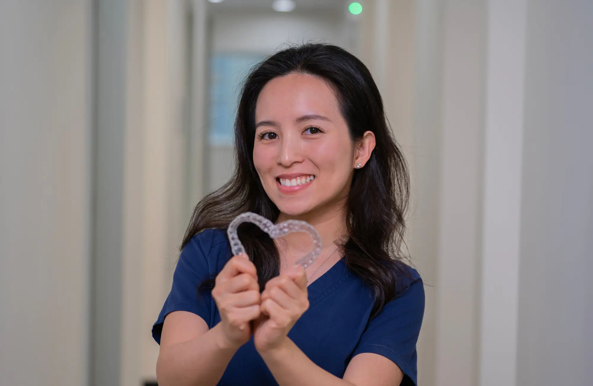 A smiling woman holds clear dental aligners shaped like a heart.