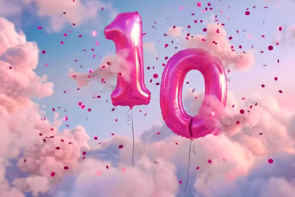 Magenta-farbene glänzende Ballons in Form der Zahl 10 vor einem pastellfarbenen Wolkenhimmel, umgeben von Konfetti – anlässlich des 10-jährigen Jubiläums.