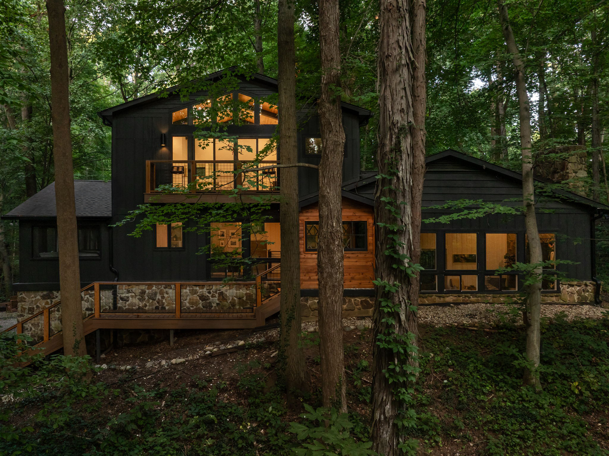 Wooded view of black modern home addition at dusk – Emergent Construction project featuring vaulted windows and natural stone foundation.