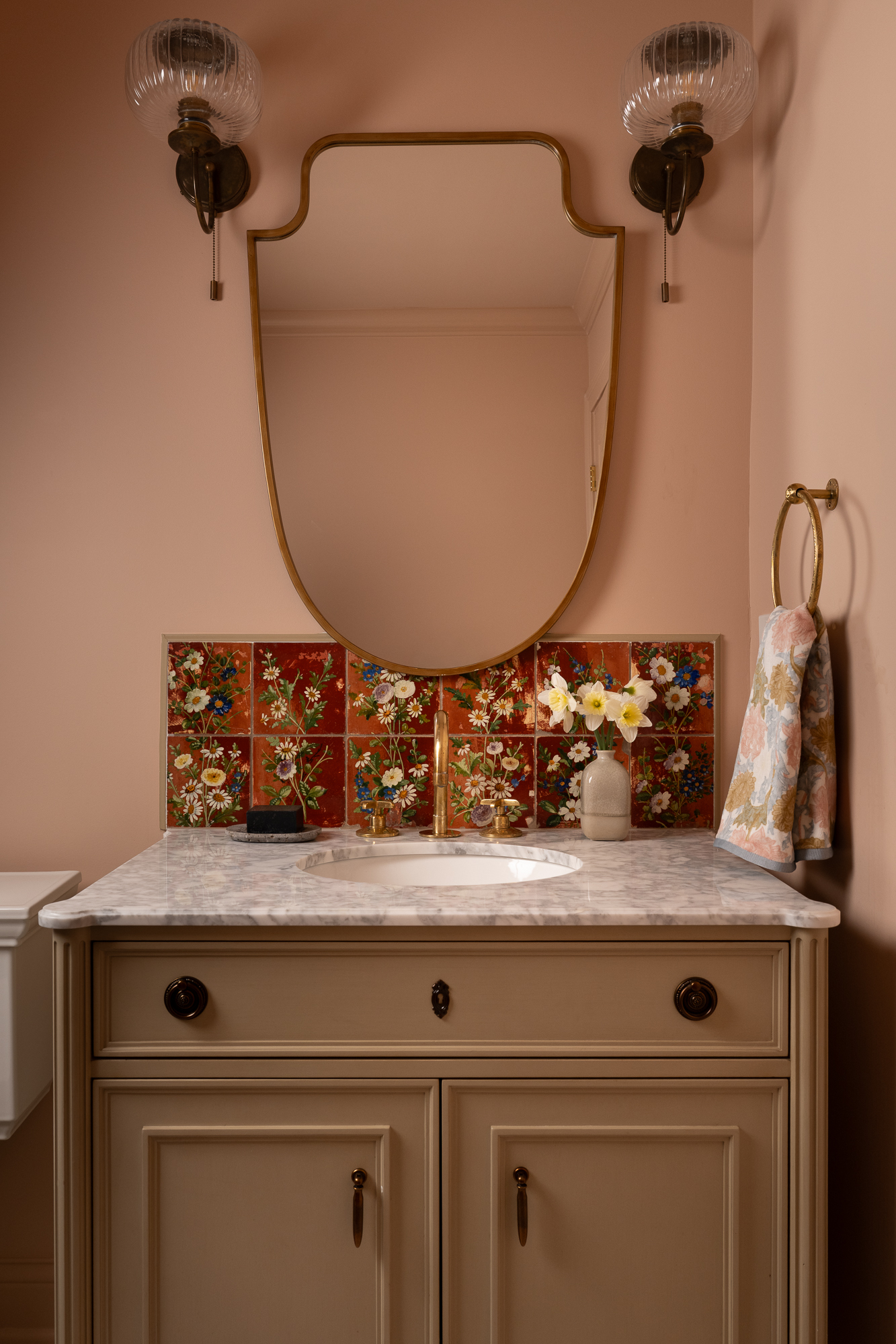 Pink powder bathroom with a cream vanity, colorful floral backsplash, brass mirror, and vintage-inspired wall sconces.