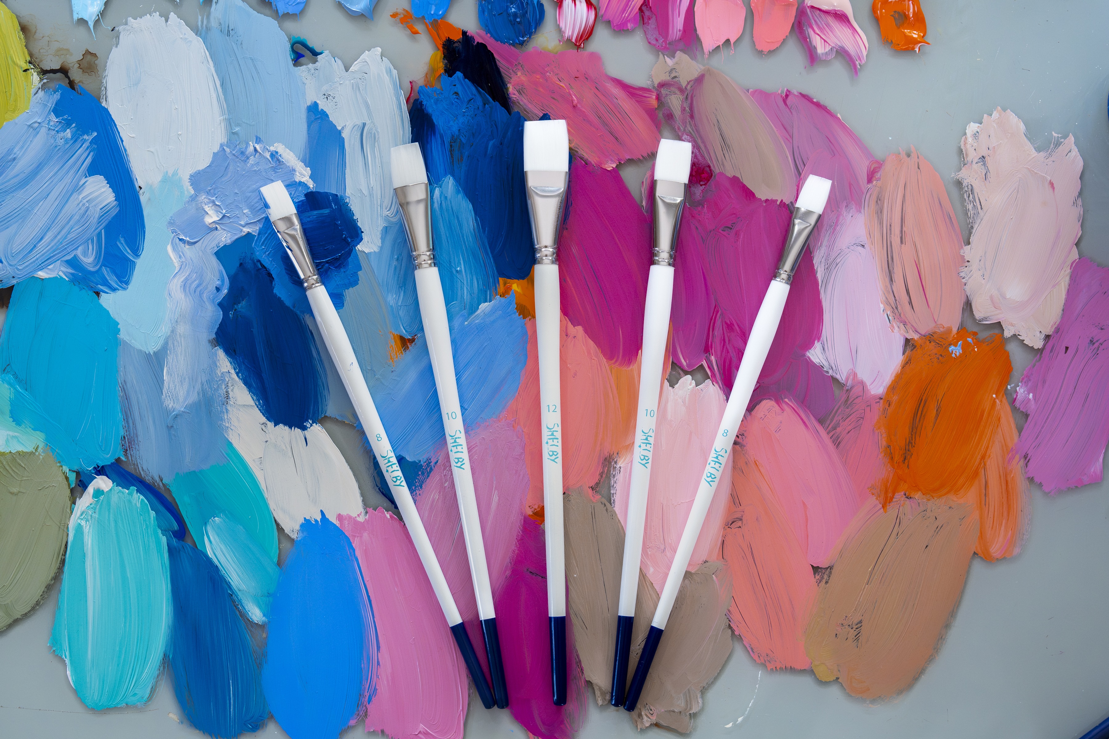 5 paint brushes lay fanned out ontop of a dried glass color palette of paints.