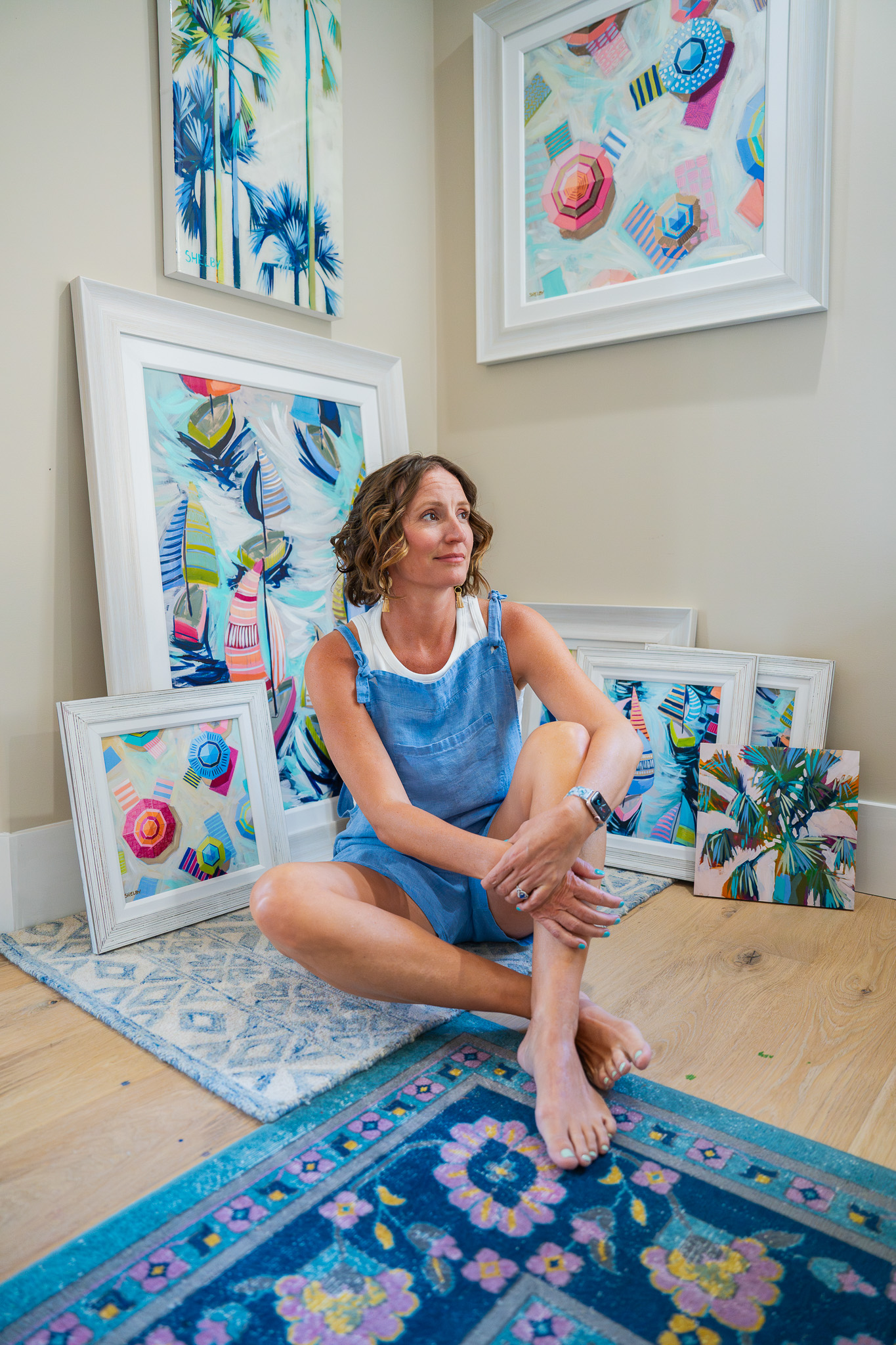 Shelby Dillon smiles while sitting cross legged in front of a group of her original paintings and prints framed and leaning against the walls in the corner that the image is shot in