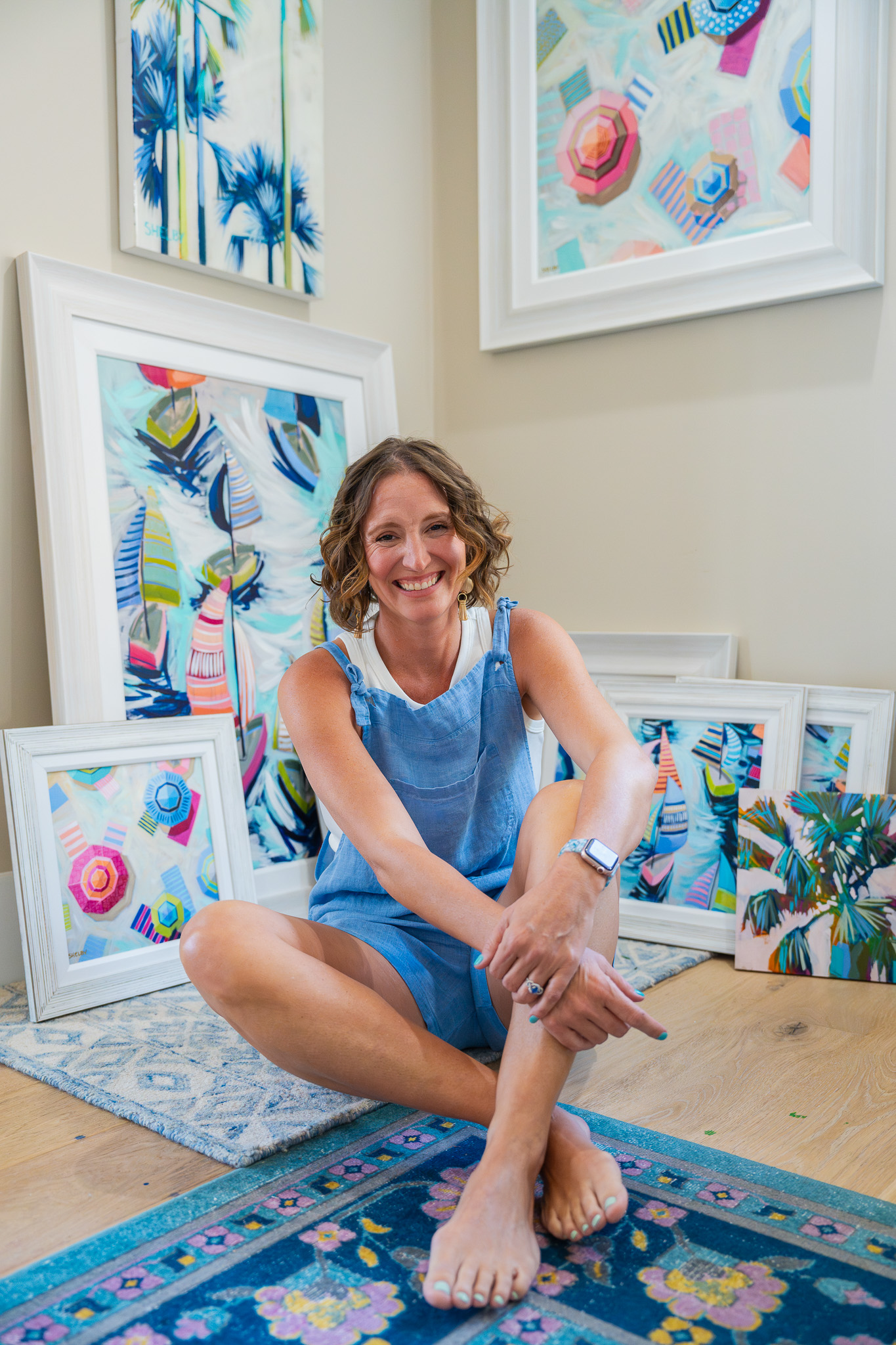 Shelby Dillon smiles while sitting cross legged in front of a group of her original paintings and prints framed and leaning against the walls in the corner that the image is shot in