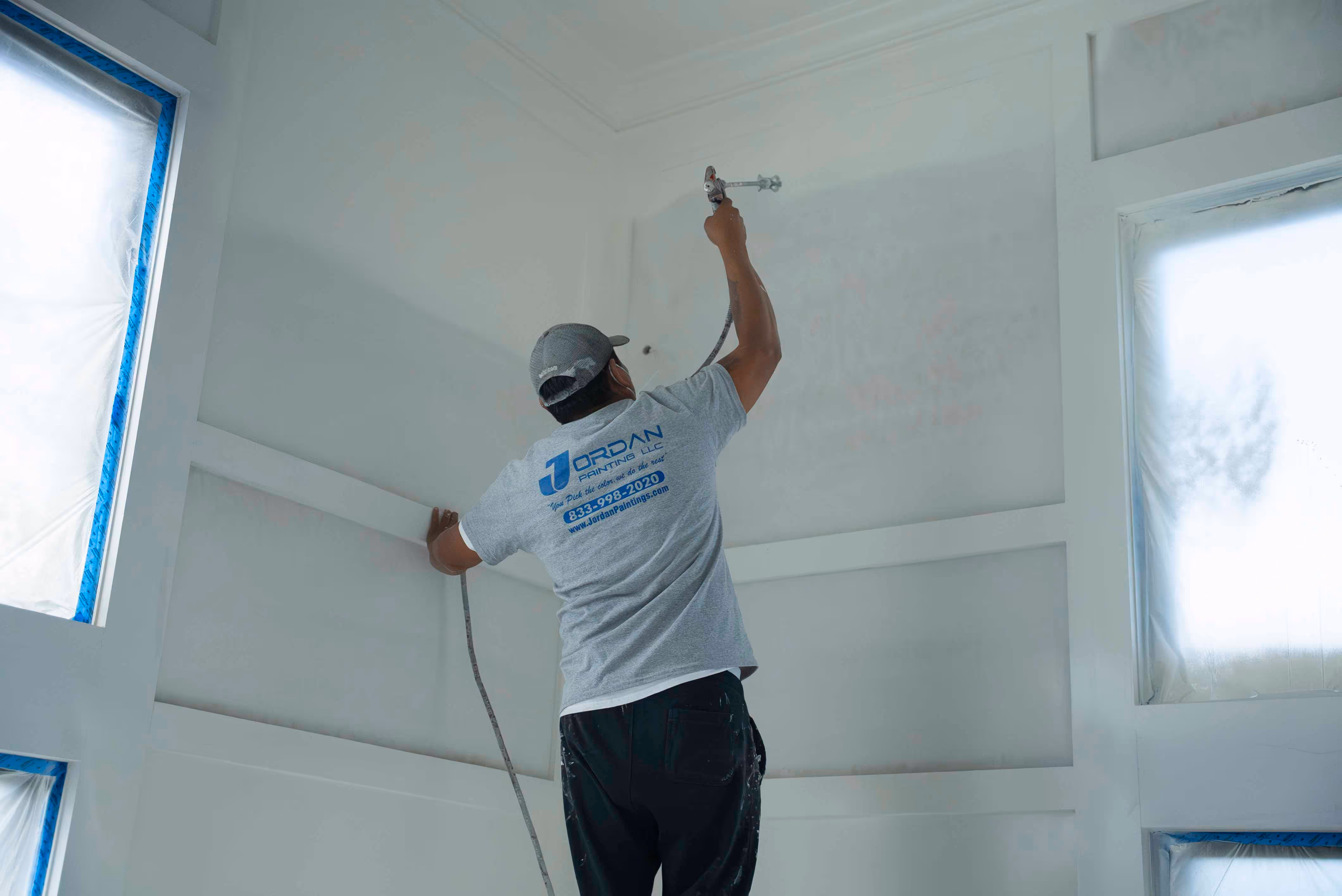 Painter in gray shirt and cap using a spray gun to paint the ceiling in a room with windows covered for protection.