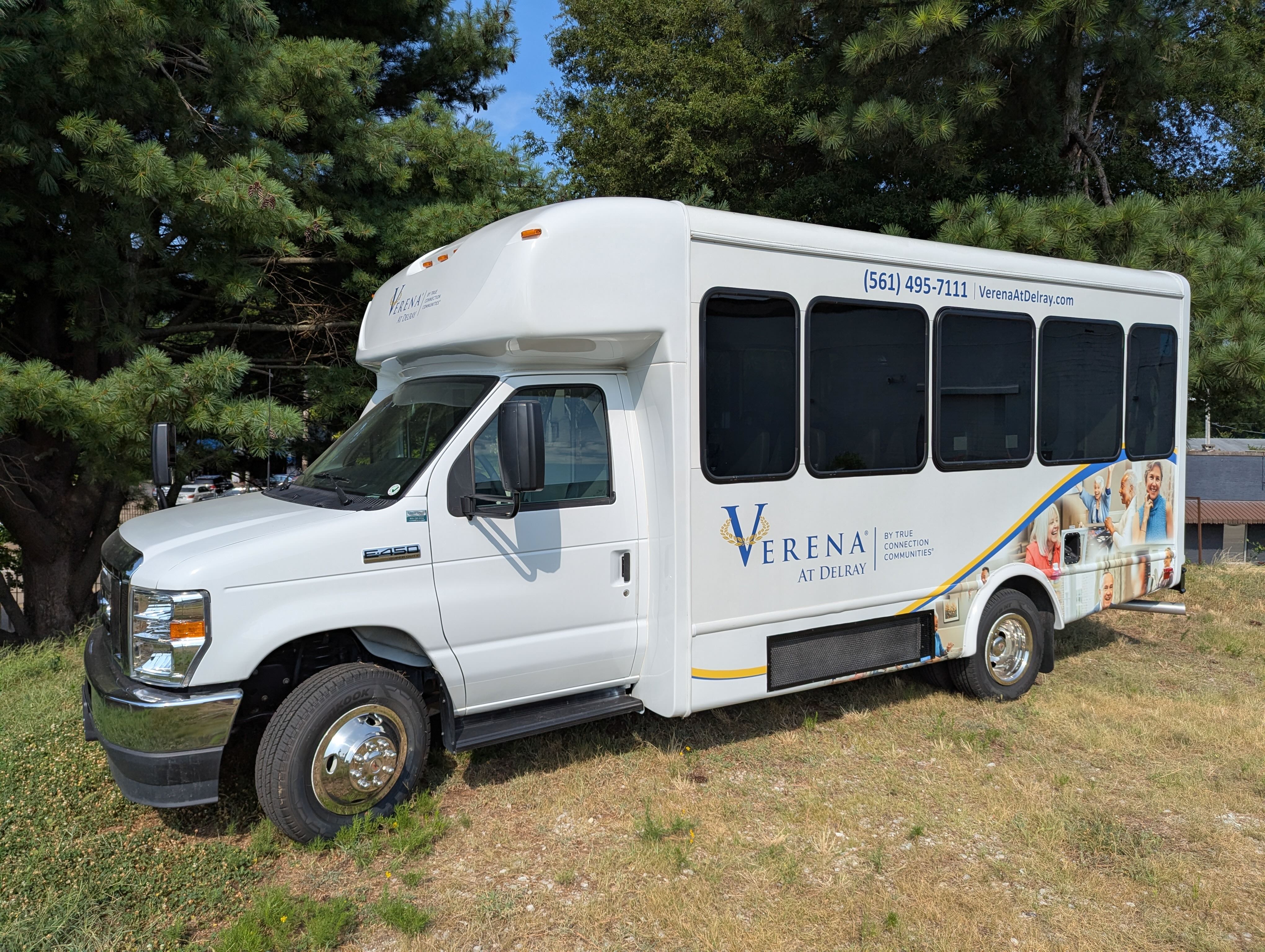 White shuttle bus parked on grass with 'Verena at Delray' branding and images of smiling elderly people on the side.