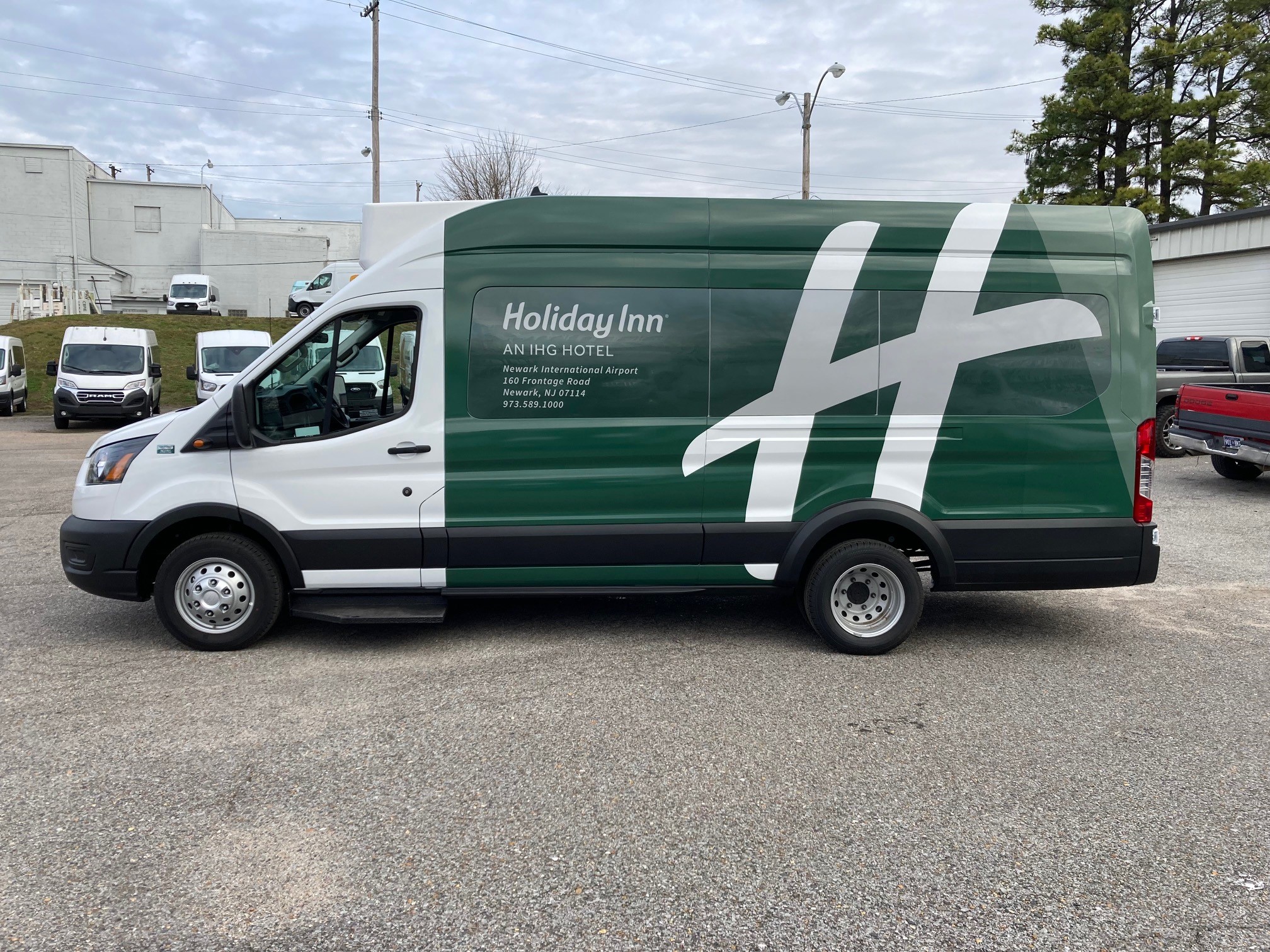 Side view of a white and green Holiday Inn hotel shuttle van parked outdoors with company branding and contact details.