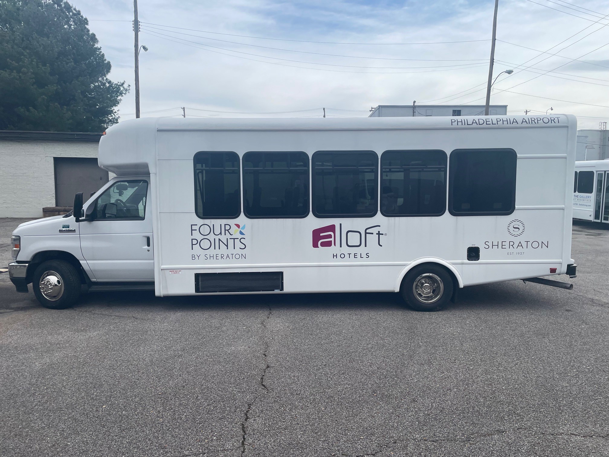 White shuttle bus parked on asphalt with logos for Four Points by Sheraton, Aloft Hotels, and Sheraton along the side and 'Philadelphia Airport' text near the roof.