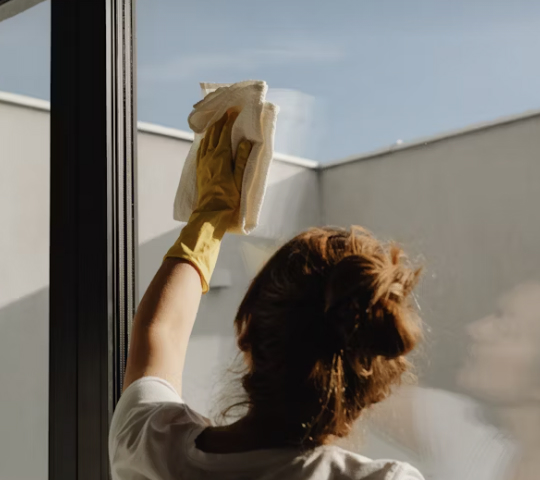 Person wearing yellow gloves cleaning a glass window with a white cloth, with sunlight and blurred outdoor background.