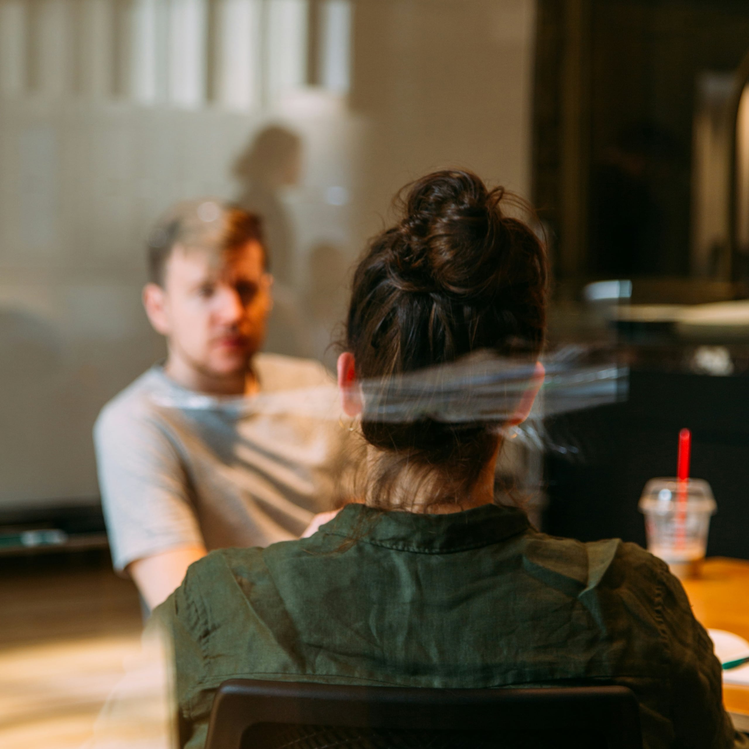 Woman with hair in a bun seated facing a man in a gray shirt, engaged in conversation at a table with a drink cup.