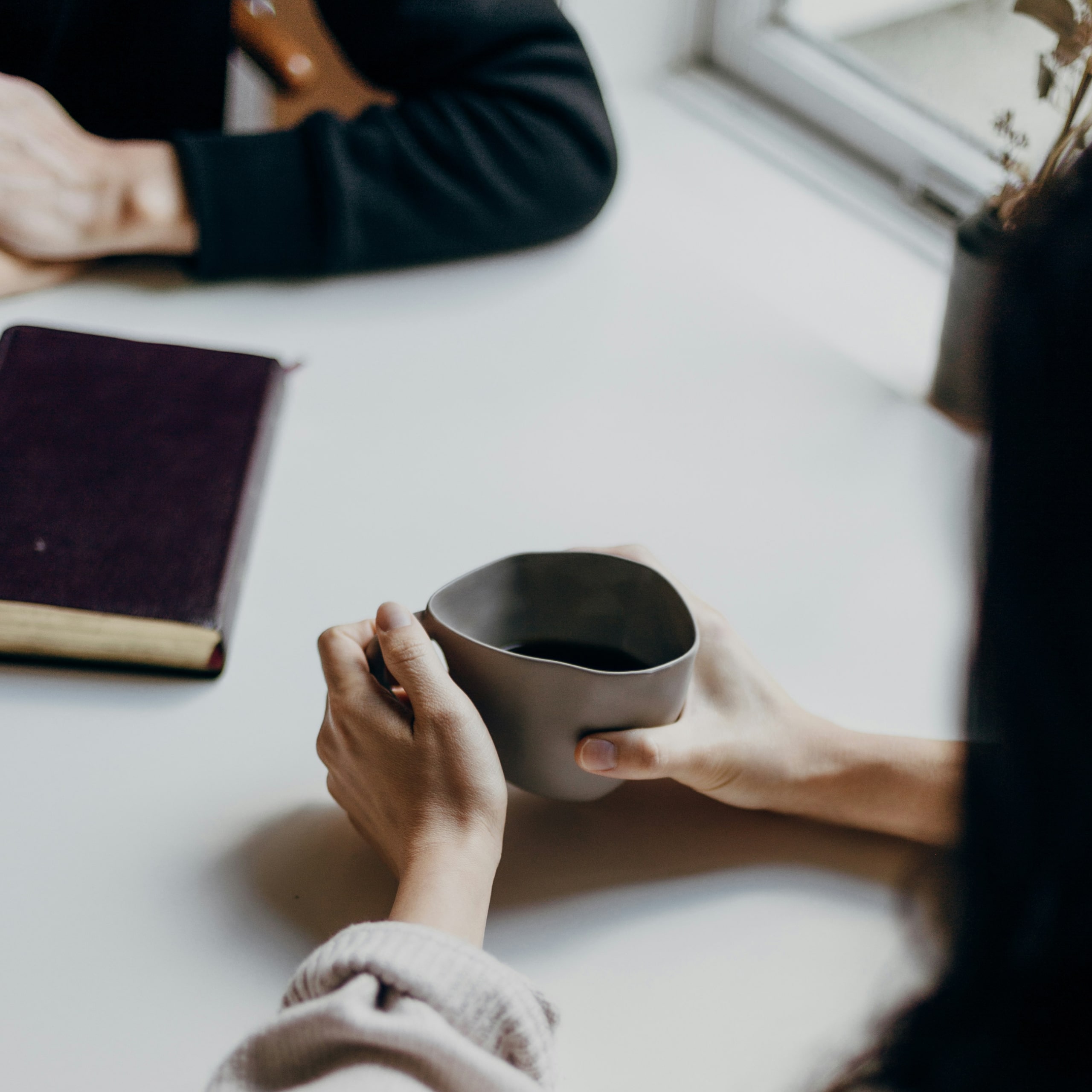 Person holding a uniquely shaped ceramic cup with both hands over a white table near another person with a closed book.