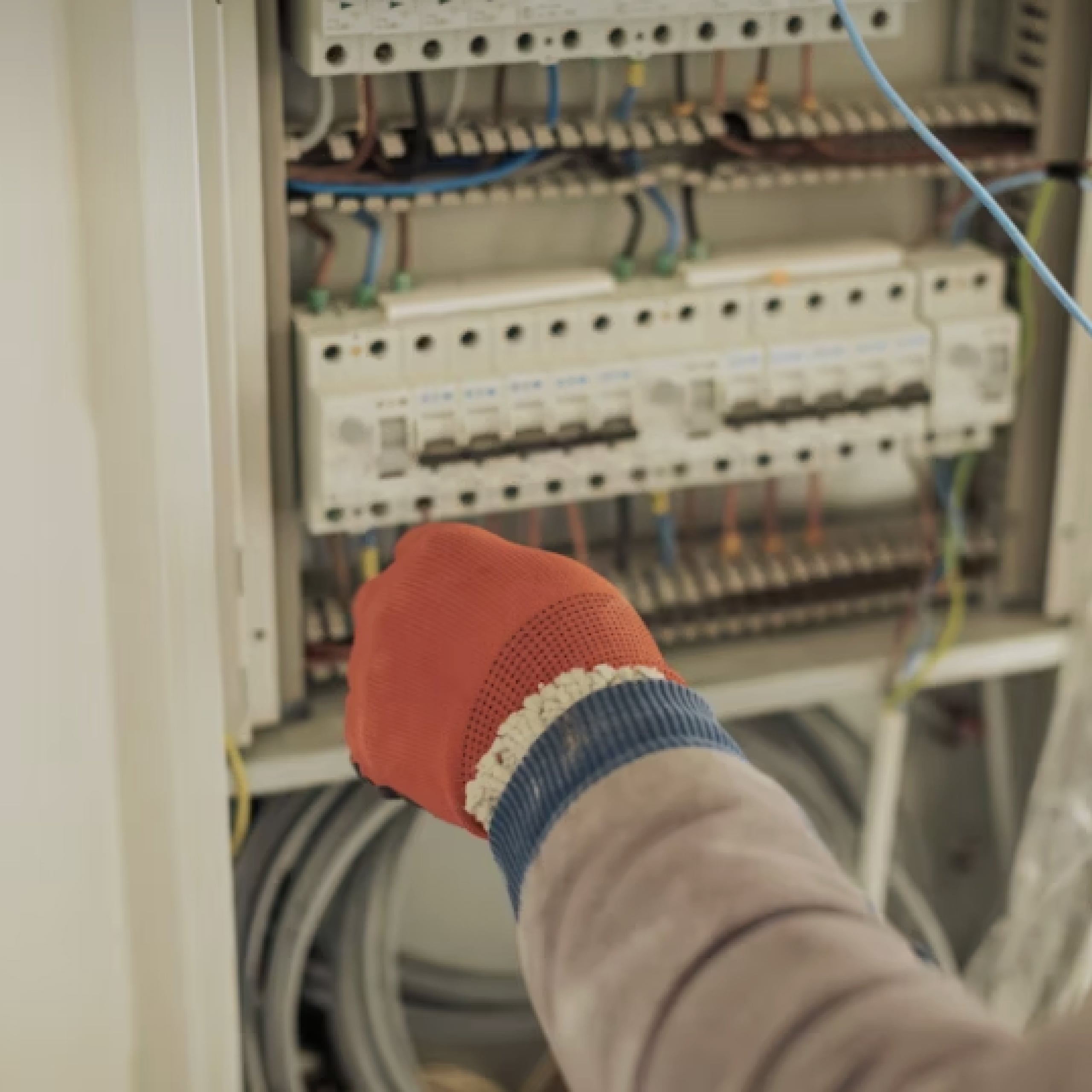 Close-up of a person wearing an orange glove working on an electrical panel with circuit breakers and exposed wiring.