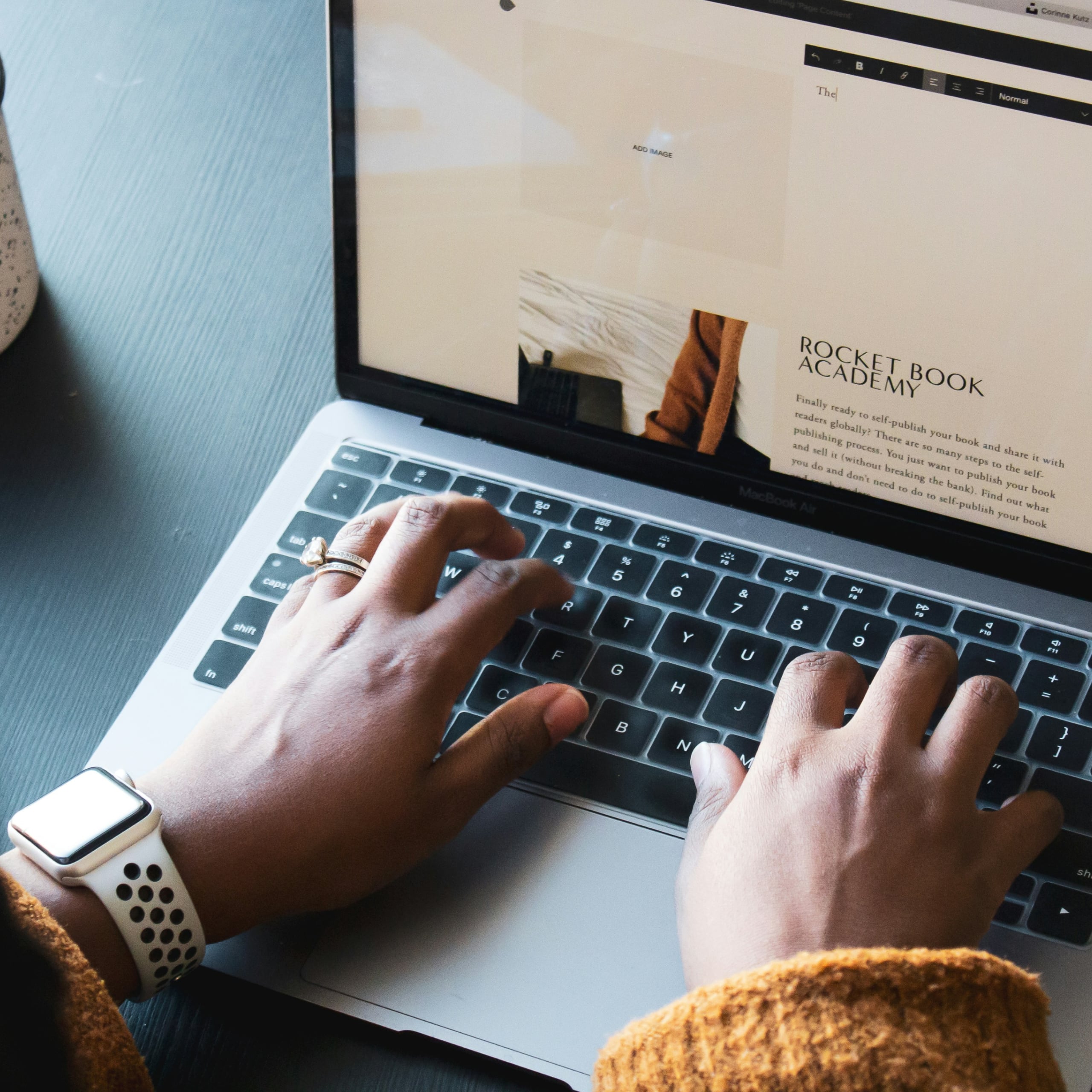 Person wearing a smartwatch and a ring typing on a MacBook Air laptop keyboard with a webpage titled 'Rocket Book Academy' visible on the screen.