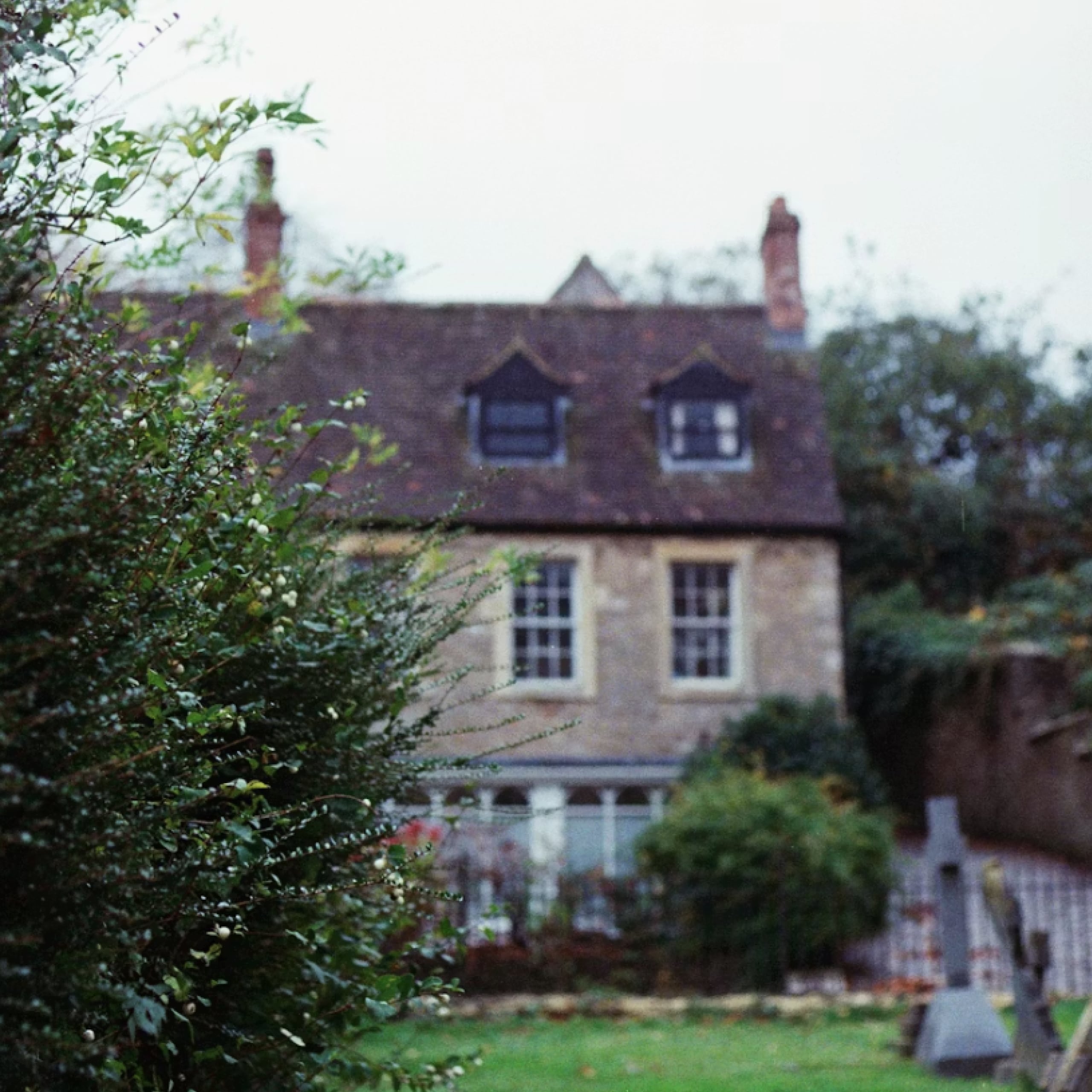 Stone house with two upper dormer windows and two front windows, partially obscured by green bushes in foreground.