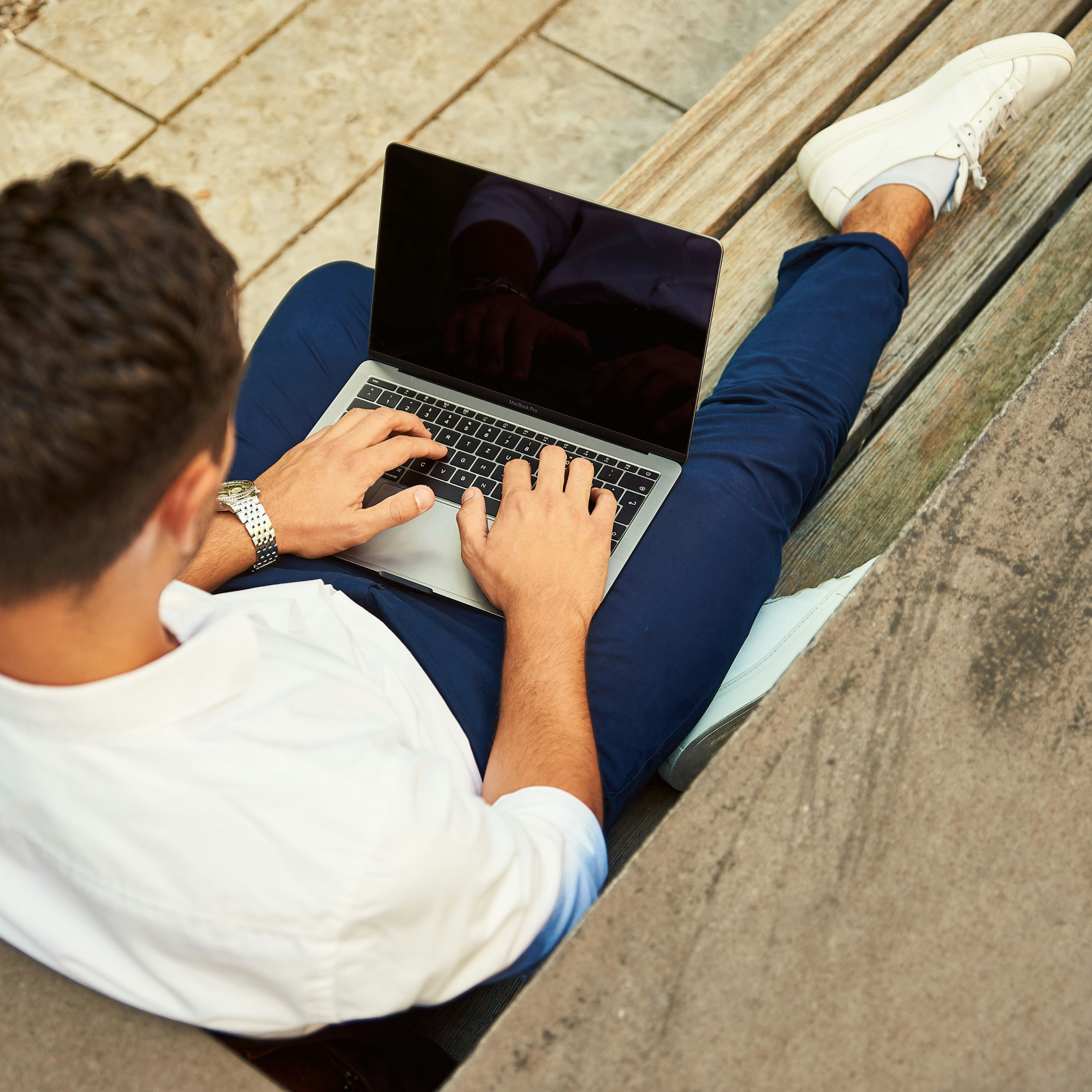 A man in a white shirt and blue pants sitting on a wooden bench typing on a laptop.