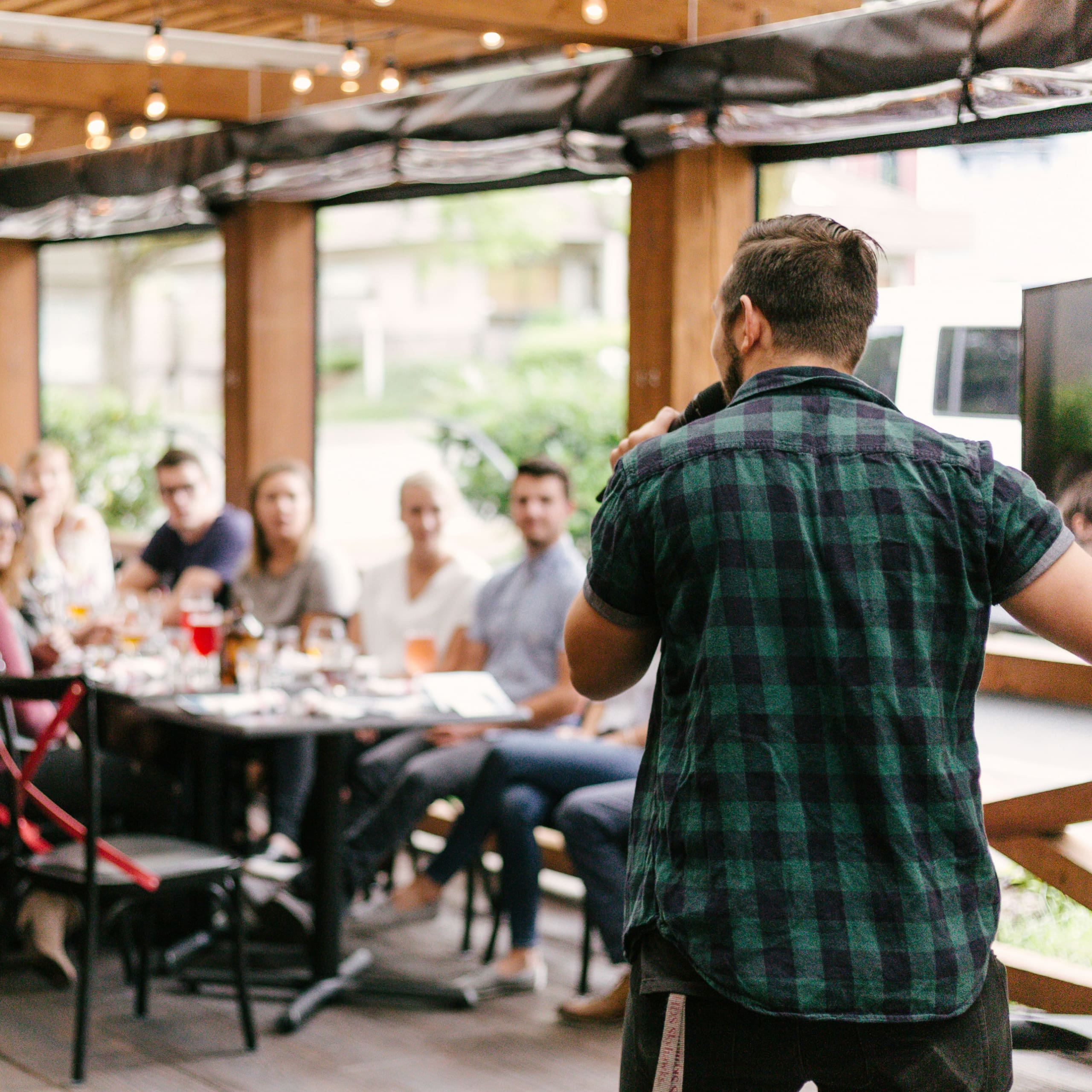 Man in a green checkered shirt speaking into a microphone to a group seated at a table in a casual indoor setting.