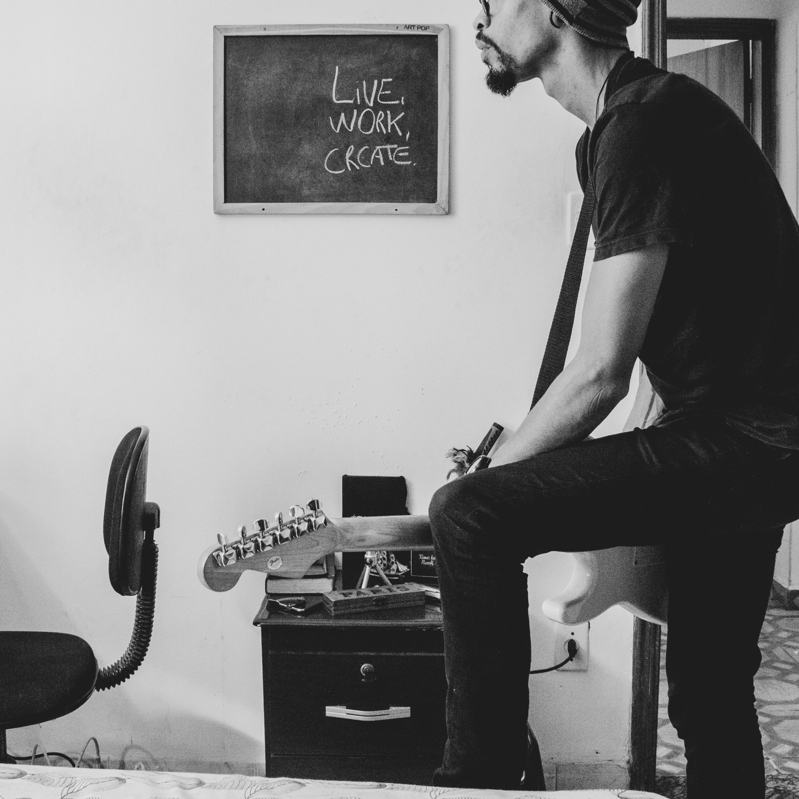 Black and white photo of a man sitting on a bed holding a guitar, with a chalkboard on the wall that reads 'Live, Work, Create.'