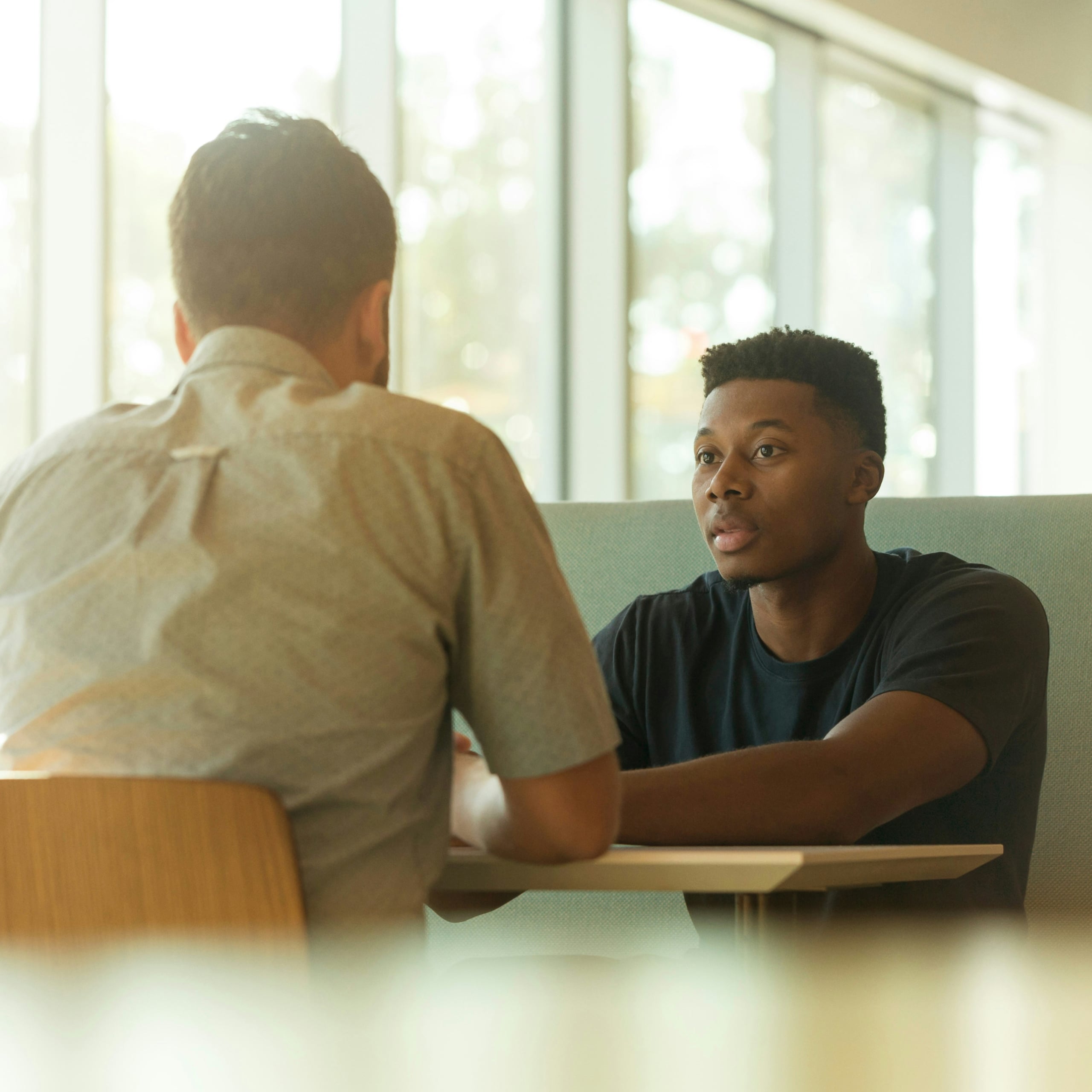 Two men sitting at a table in a bright room having a serious conversation.