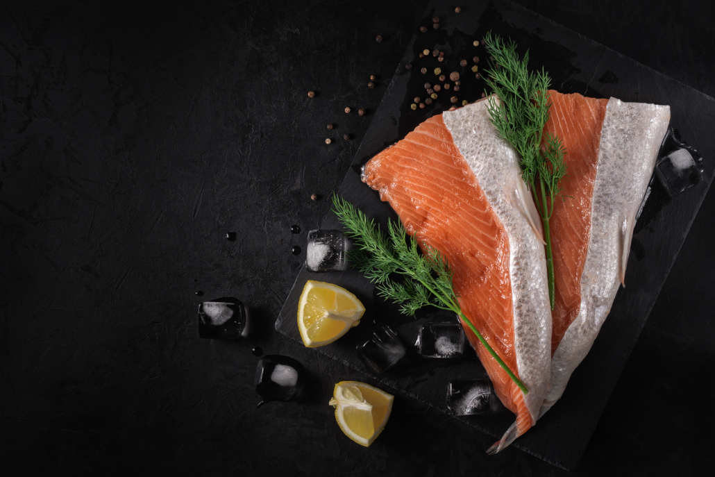 sliced fresh raw salmon lying on a stone cutting board with lemon wedges, green dill, spices and ice cubes on a black textured concrete background. top view. beautiful sea food concept with copy space