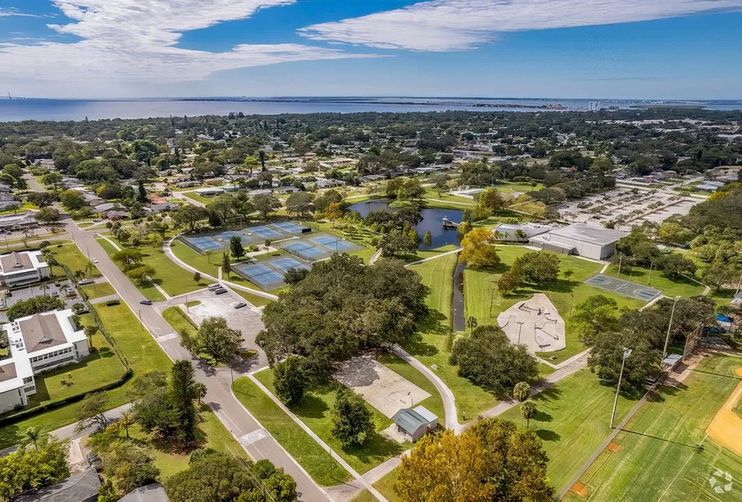 Aerial view of a suburban area featuring tennis courts, a playground, a baseball field, and a lake surrounded by greenery. Residential houses and buildings are visible in the background with a body of water in the distance under a partly cloudy sky.