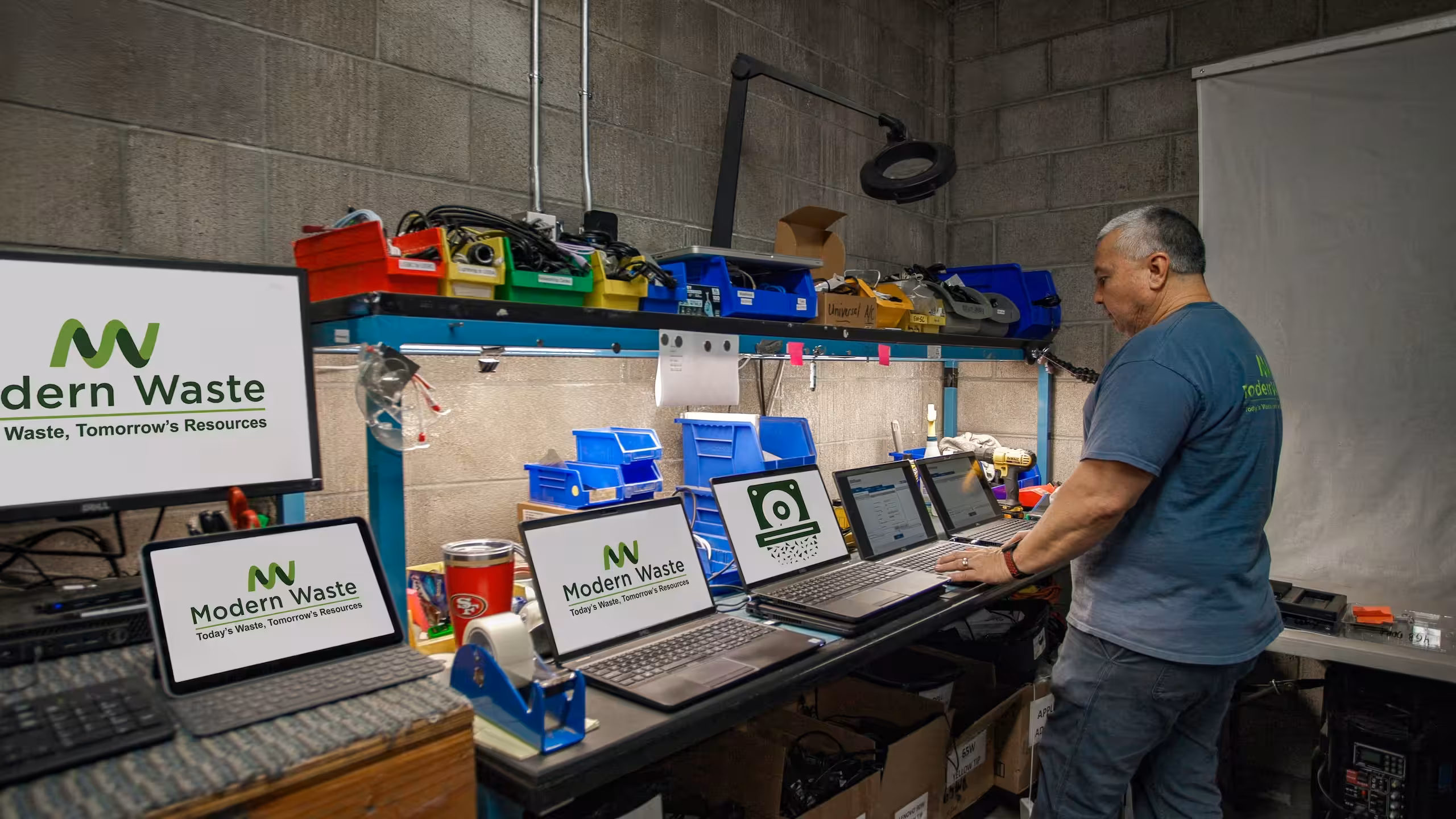 Man in a workshop standing at a bench with multiple laptops and screens displaying Modern Waste logos and data.