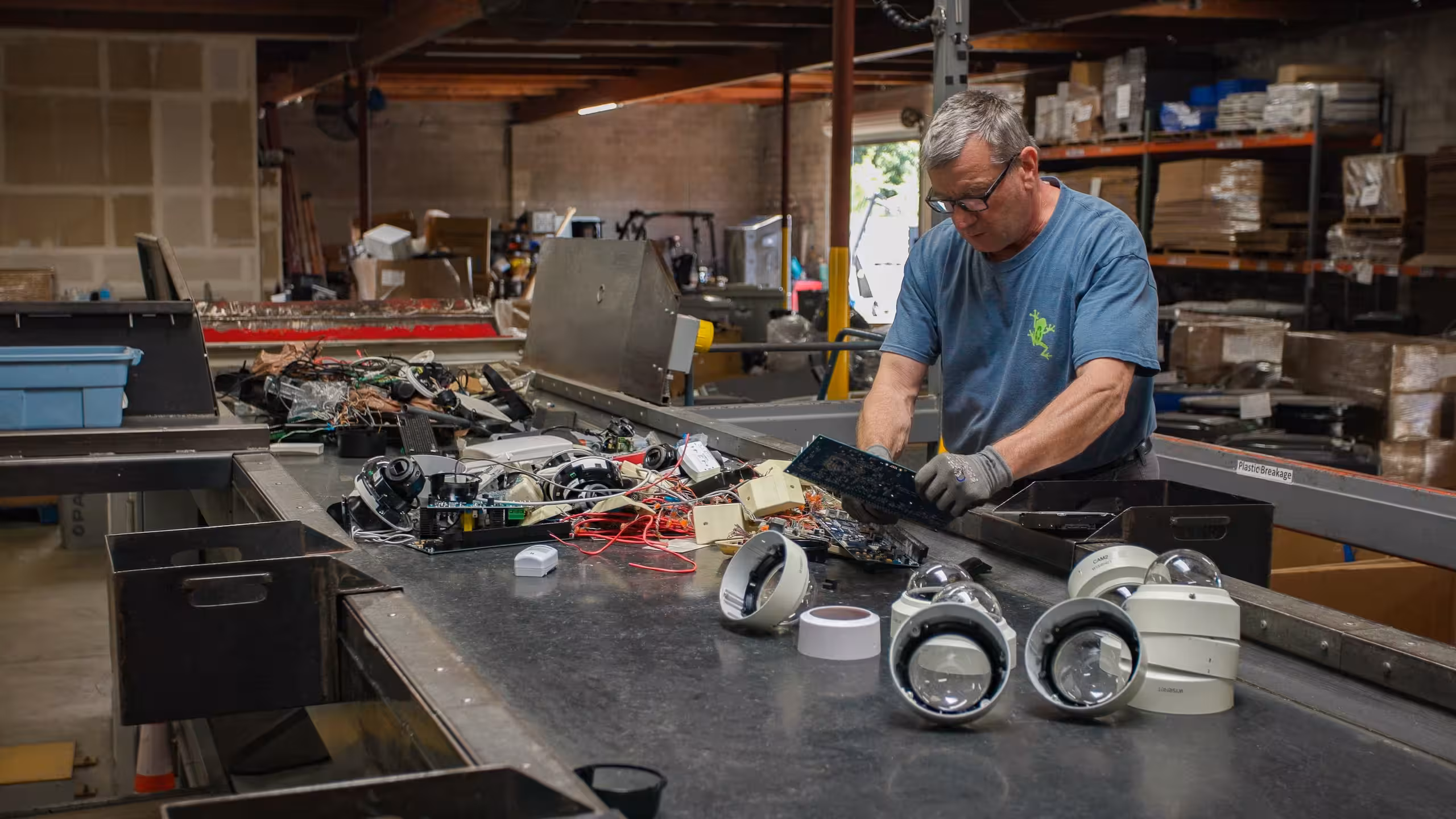 Man in blue shirt working with electronic circuit boards and security camera parts on a large conveyor table in an industrial warehouse.