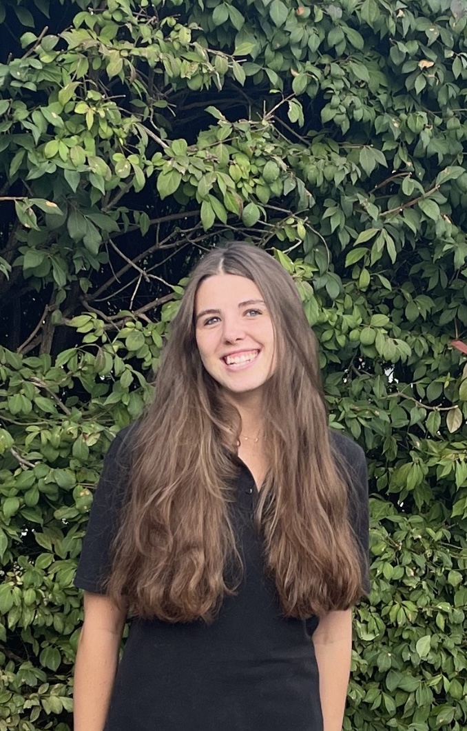 Young woman with long brown hair poses in a lush green outdoor landscape setting.