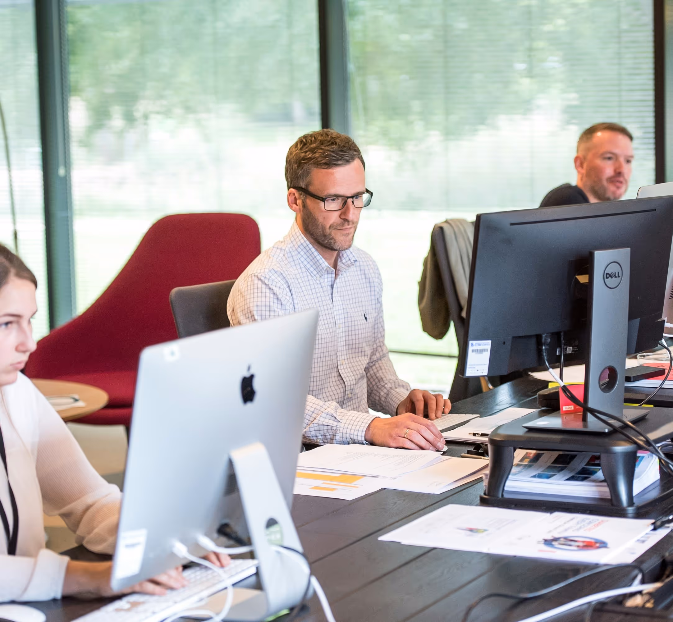Three people working at computers in a modern office with large windows. A man in glasses and a checkered shirt sits at a desk with a Dell monitor and MacBook, smiling while working. A woman with a white laptop is visible on the left, and another man is seated in the background. The office has contemporary furniture including a red accent chair."
This alt text describes the key elements: the number of people, their positioning, the technology they're using, the office environment, and notable furniture pieces.