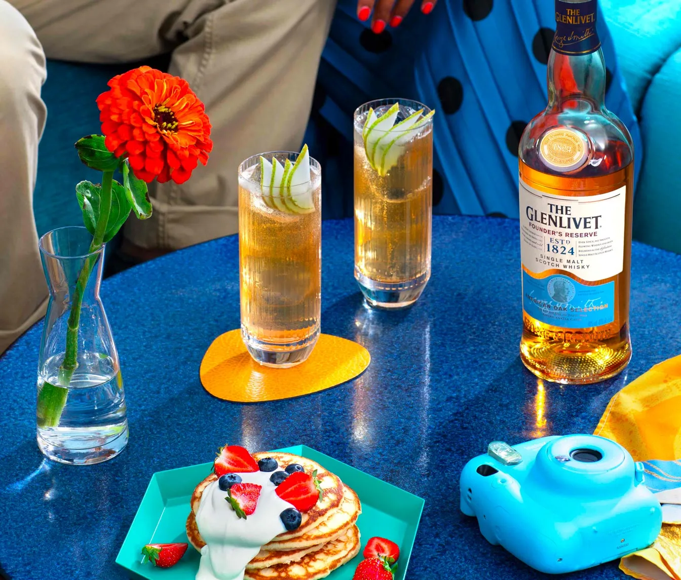 Blue table with a vase holding a bright orange flower, two tall glasses of amber-colored drinks garnished with thinly sliced green apple, a bottle of The Glenlivet Scotch whisky, a blue instant camera, and a turquoise plate with pancakes topped with yogurt, strawberries, and blueberries.