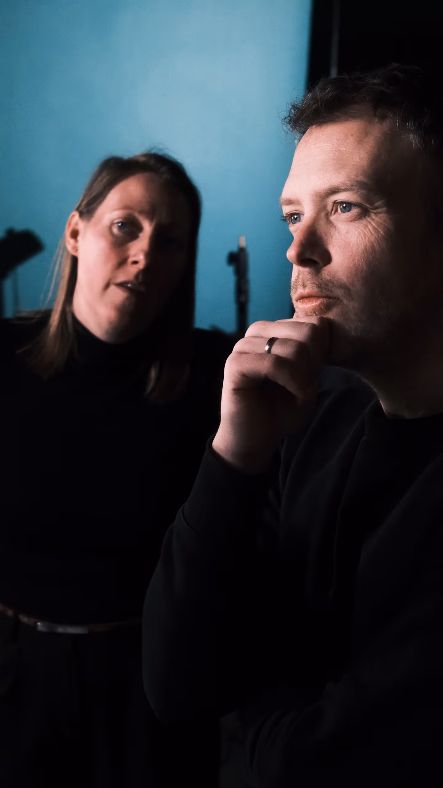 Man met een zwarte trui denkt diep na met zijn hand aan zijn kin, vrouw op de achtergrond tegen een blauwe muur.