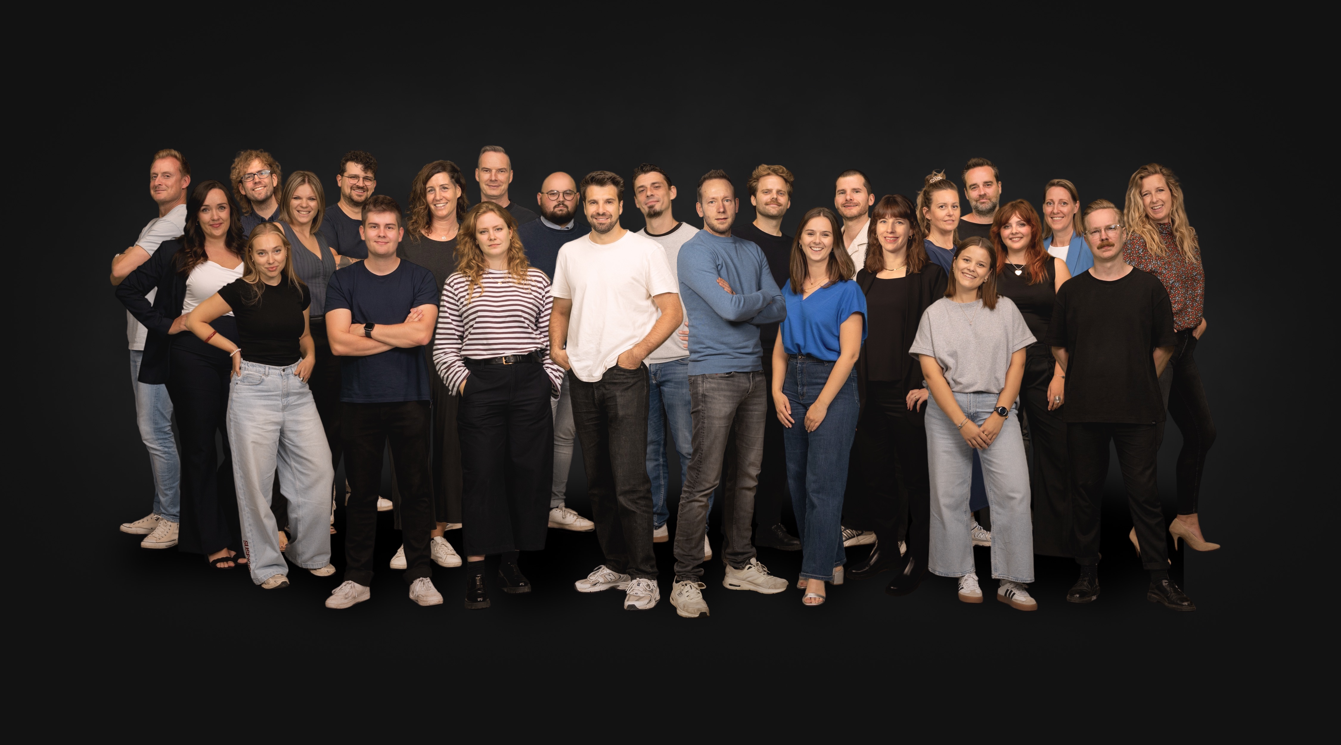 Groepsfoto van het team van Medialife die voor een zwarte achtergrond staan, gekleed in casual en zakelijke kleding.