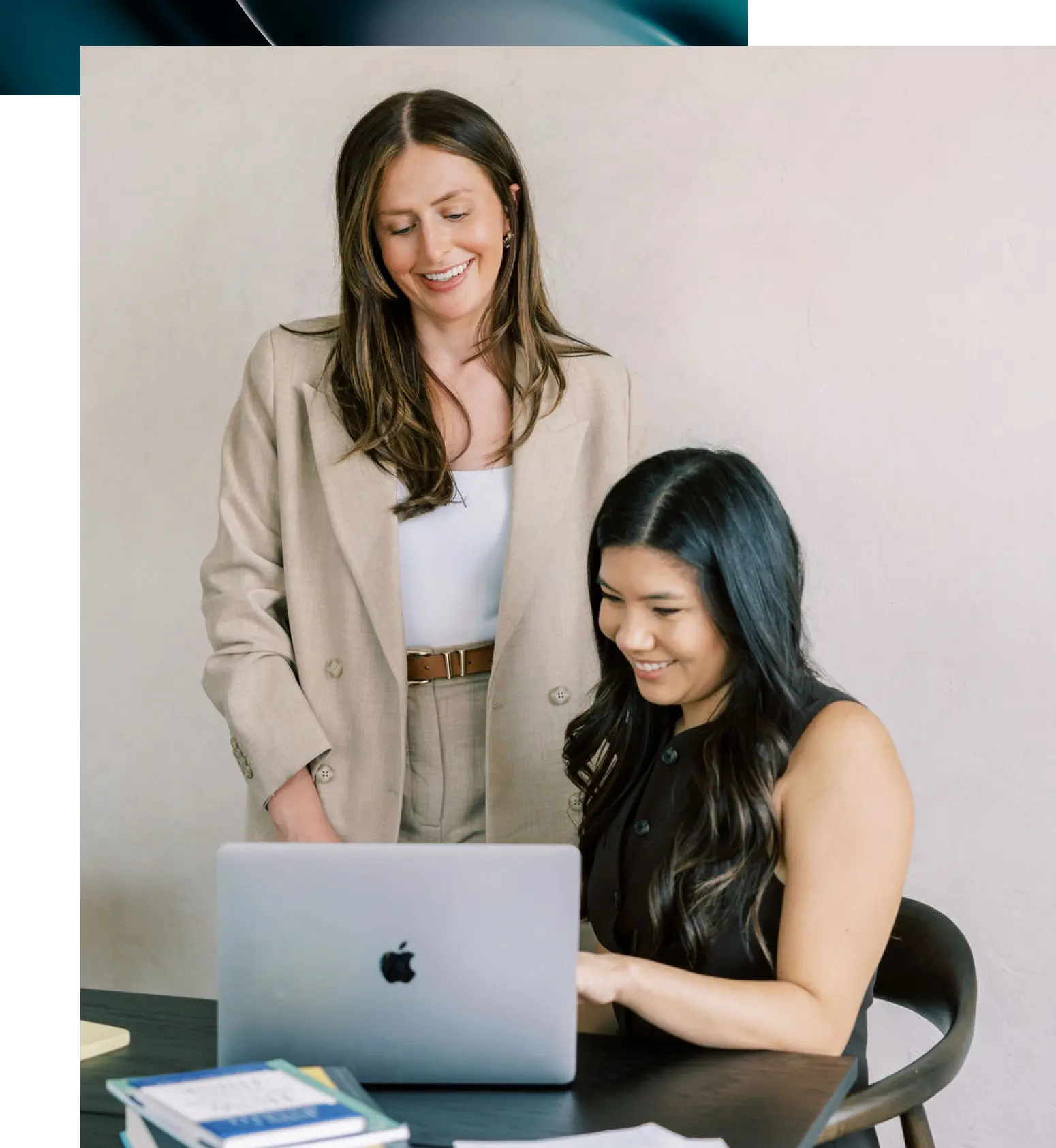 Two women sitting at a white table, smiling and working together with a notebook and laptop in a bright, modern room.